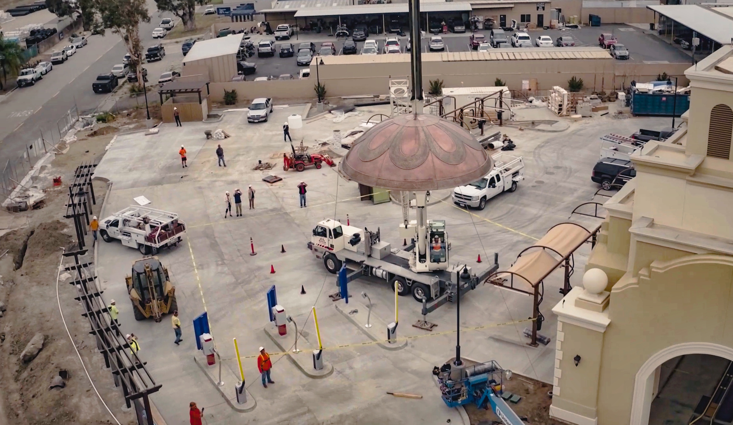 Construction workers and equipment working on a car wash installation, with cars parked in the background, a decorative dome structure, and construction vehicles and tools around.