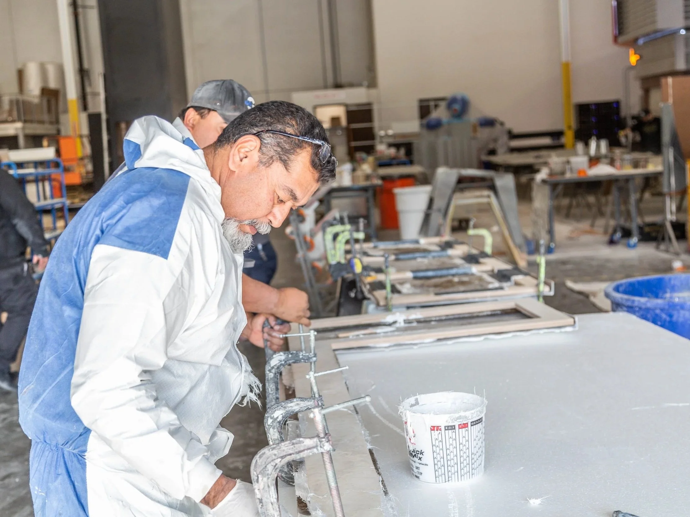 Two men working in a factory or workshop, wearing protective clothing and glasses, focused on the assembly or construction of items on a table, with tools and materials around them.