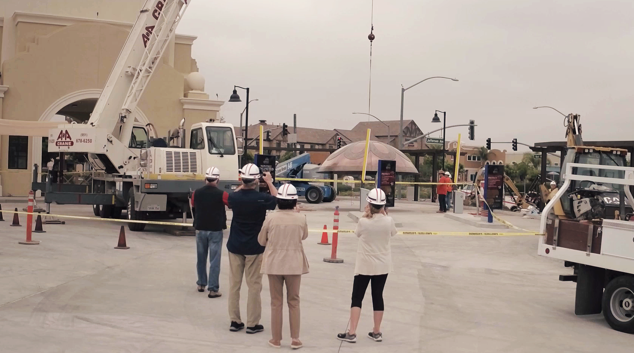 People wearing safety helmets watching construction work with cranes and equipment in an urban area, police tape cordoning off the area, under a cloudy sky.