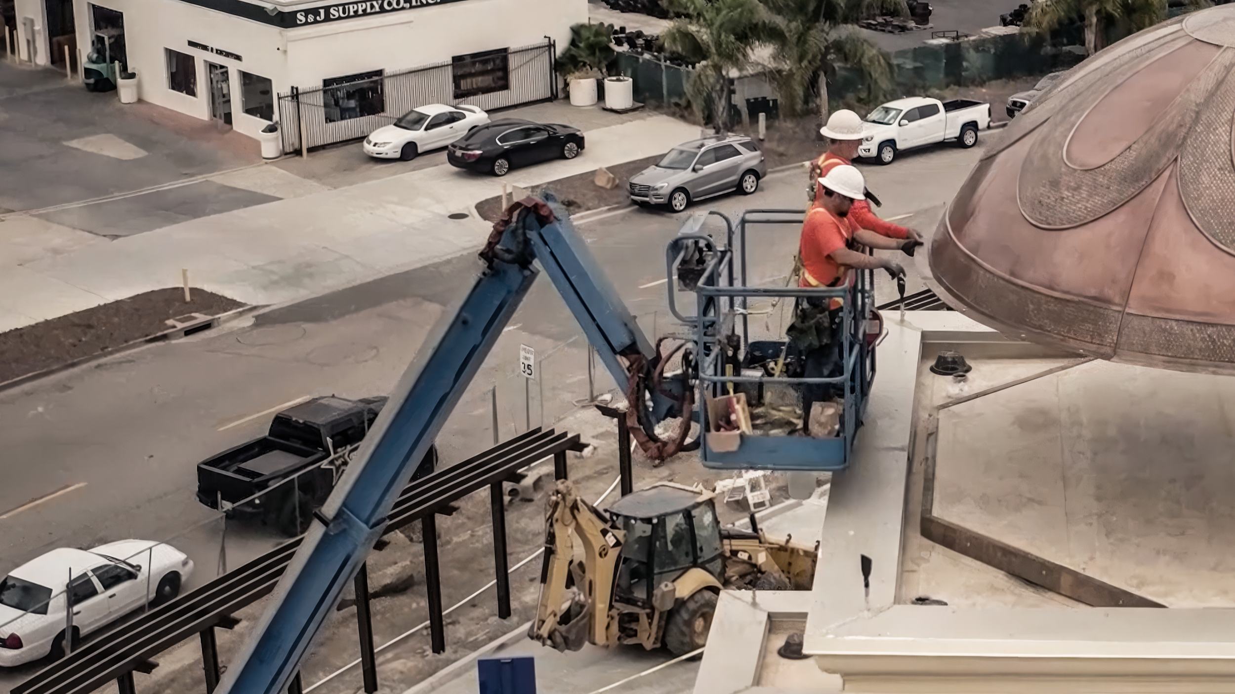 Construction workers in orange shirts and white helmets operating a lift to work on a building with a copper-colored dome.