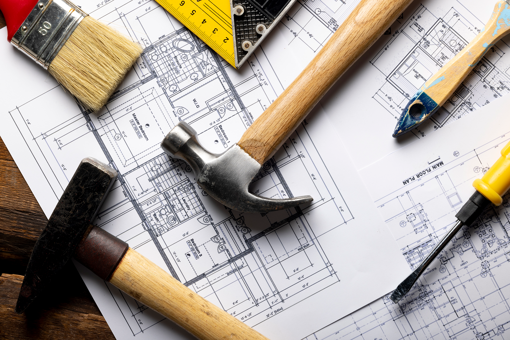 Construction tools including a paintbrush, hammer, screwdriver, and measuring tape laid on Top of architectural blueprints.