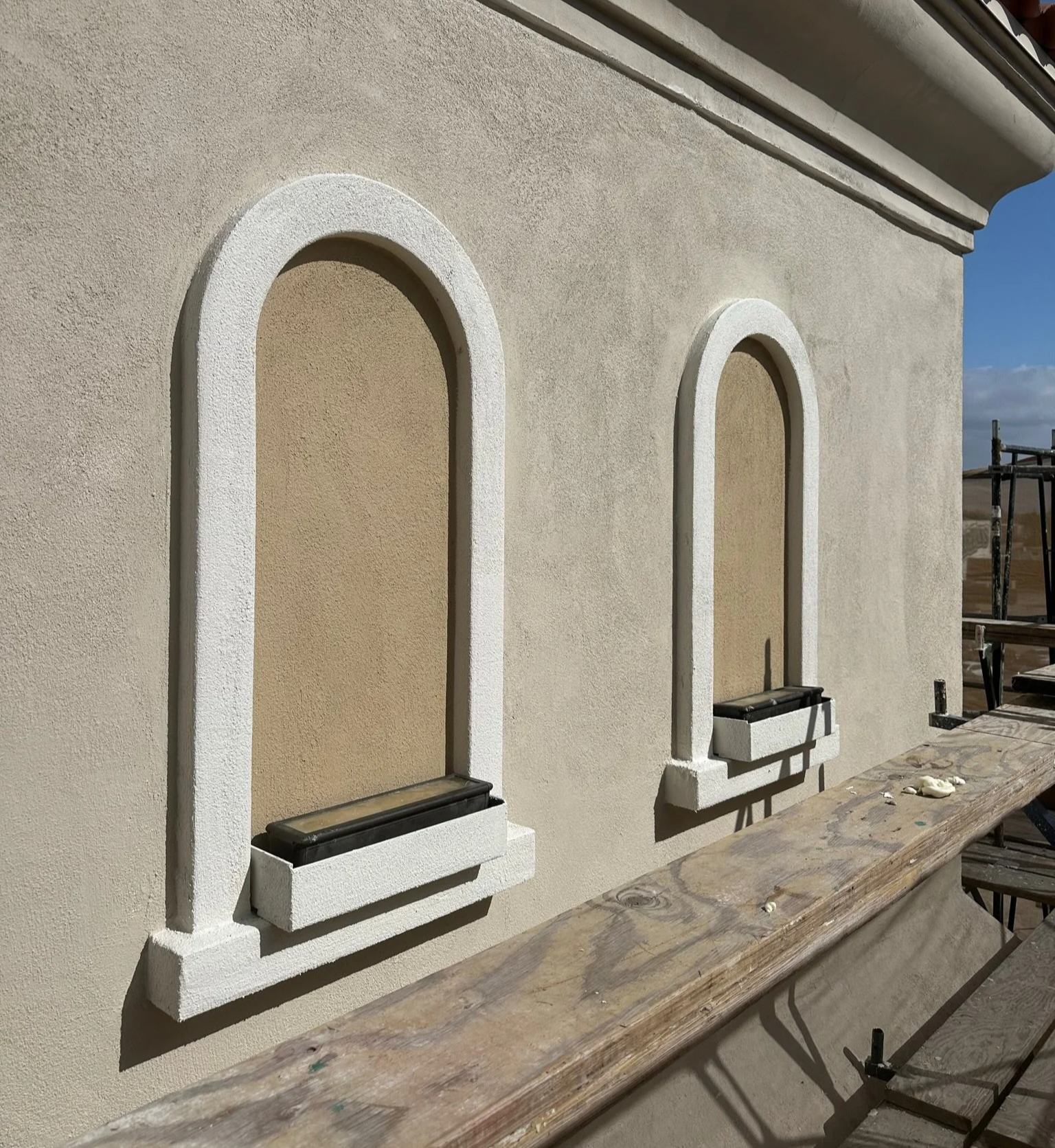 Construction site with a beige stucco wall featuring three arched window openings with white trim and window planters, a wooden workbench, and scaffolding outside.