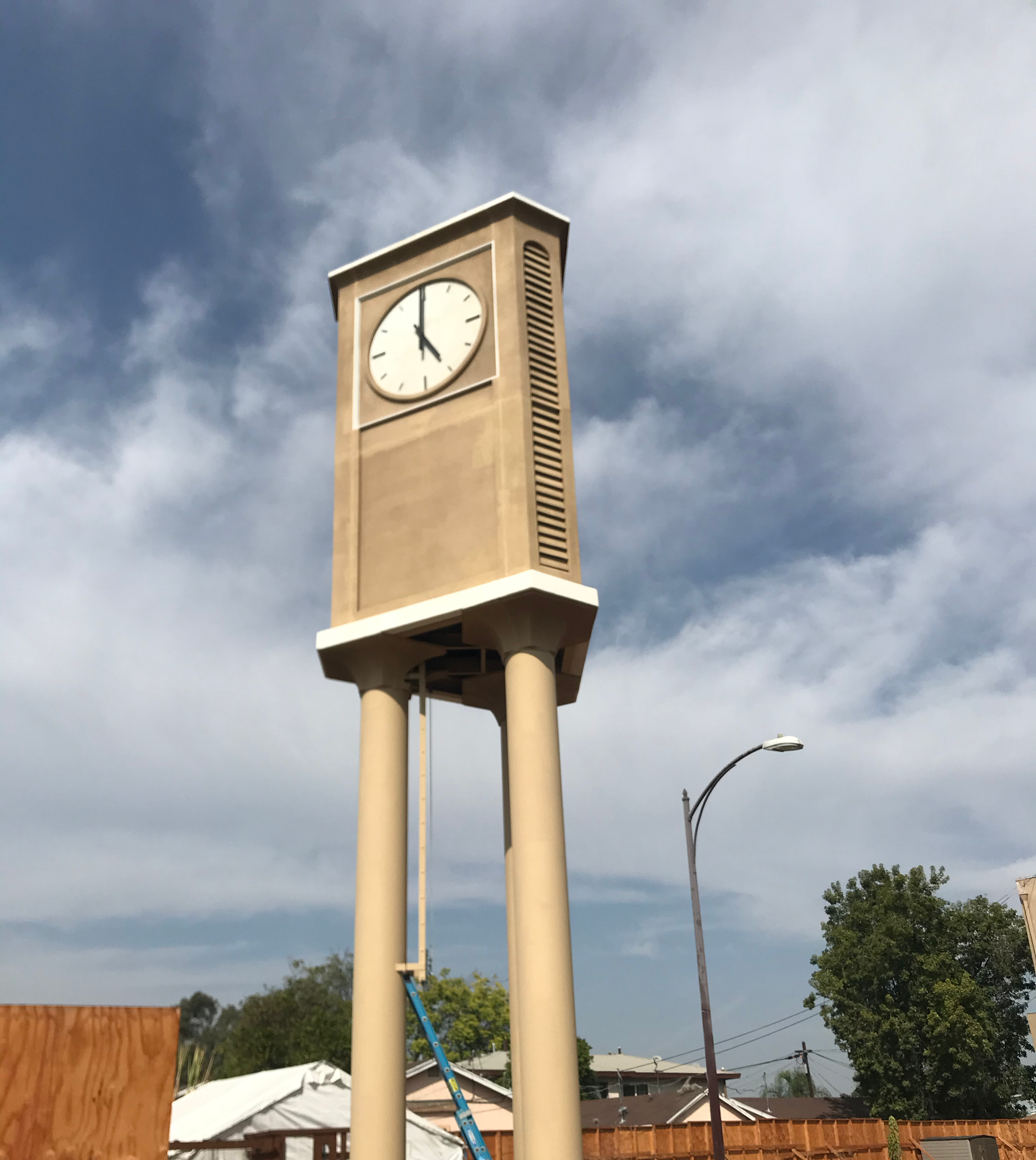 A clock tower with a white clock face showing around 4:56, beige structure, and beige columns, set against a cloudy sky with a streetlamp and residential houses in the background.