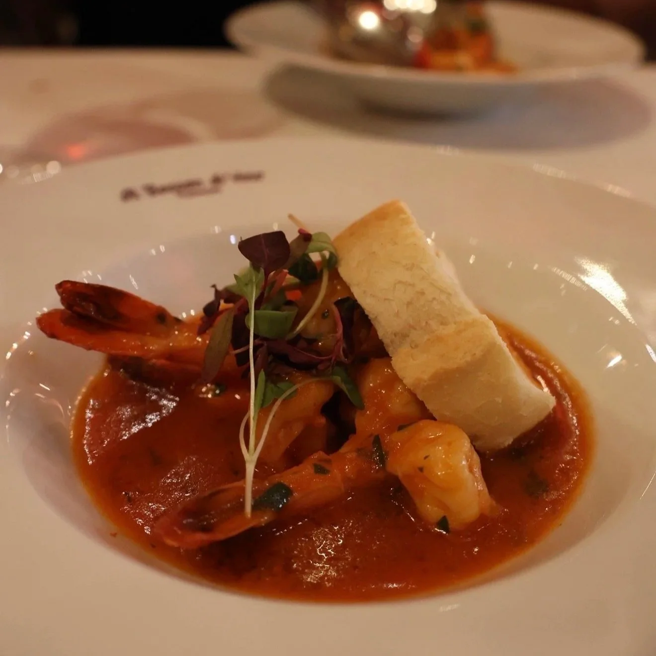 Close-up of a seafood dish featuring shrimp and a slice of baguette on a bed of tomato-based sauce, garnished with microgreens, served on a white plate.