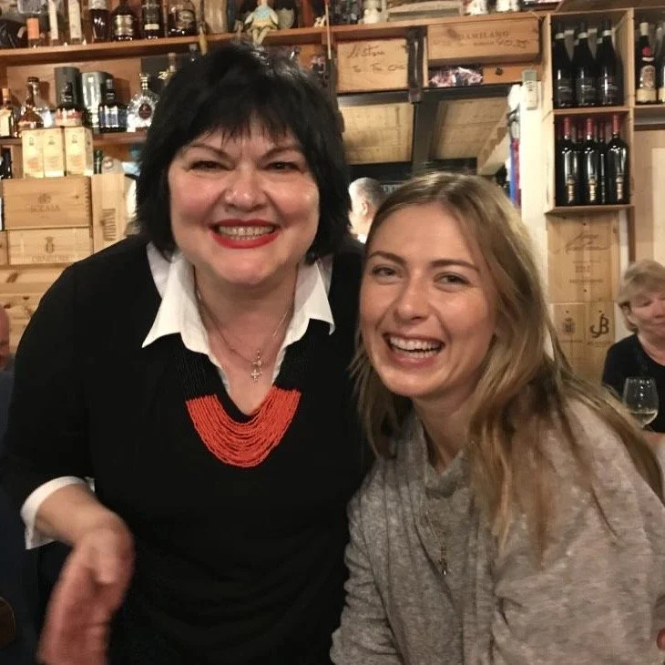 Two women smiling and posing for a photo in a cozy, rustic restaurant with wooden shelves and wine bottles in the background.