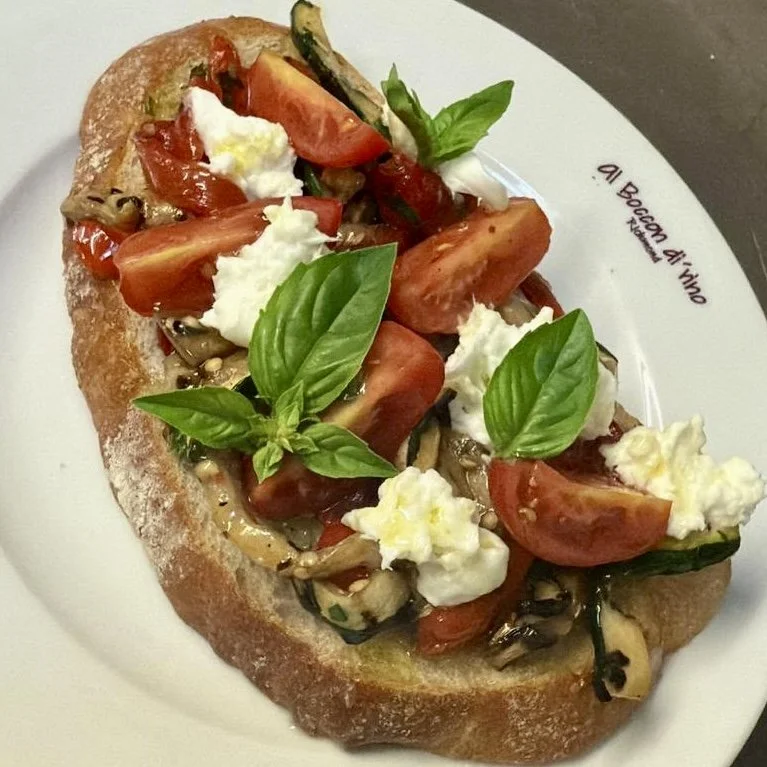 Open-faced toast topped with sliced tomatoes, fresh basil leaves, dollops of ricotta cheese, and sautéed vegetables on a white plate.