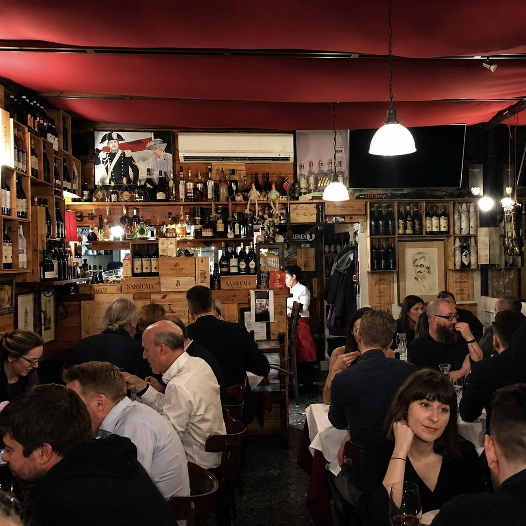 Inside a busy restaurant with diners seated at tables, wooden shelves filled with wine bottles, decorative artwork, and hanging pendant lights.