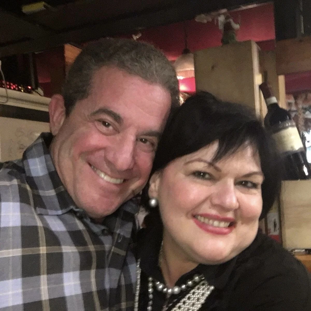 A smiling man and woman posing closely together in a dimly lit restaurant or bar, with a champagne bottle in the background.