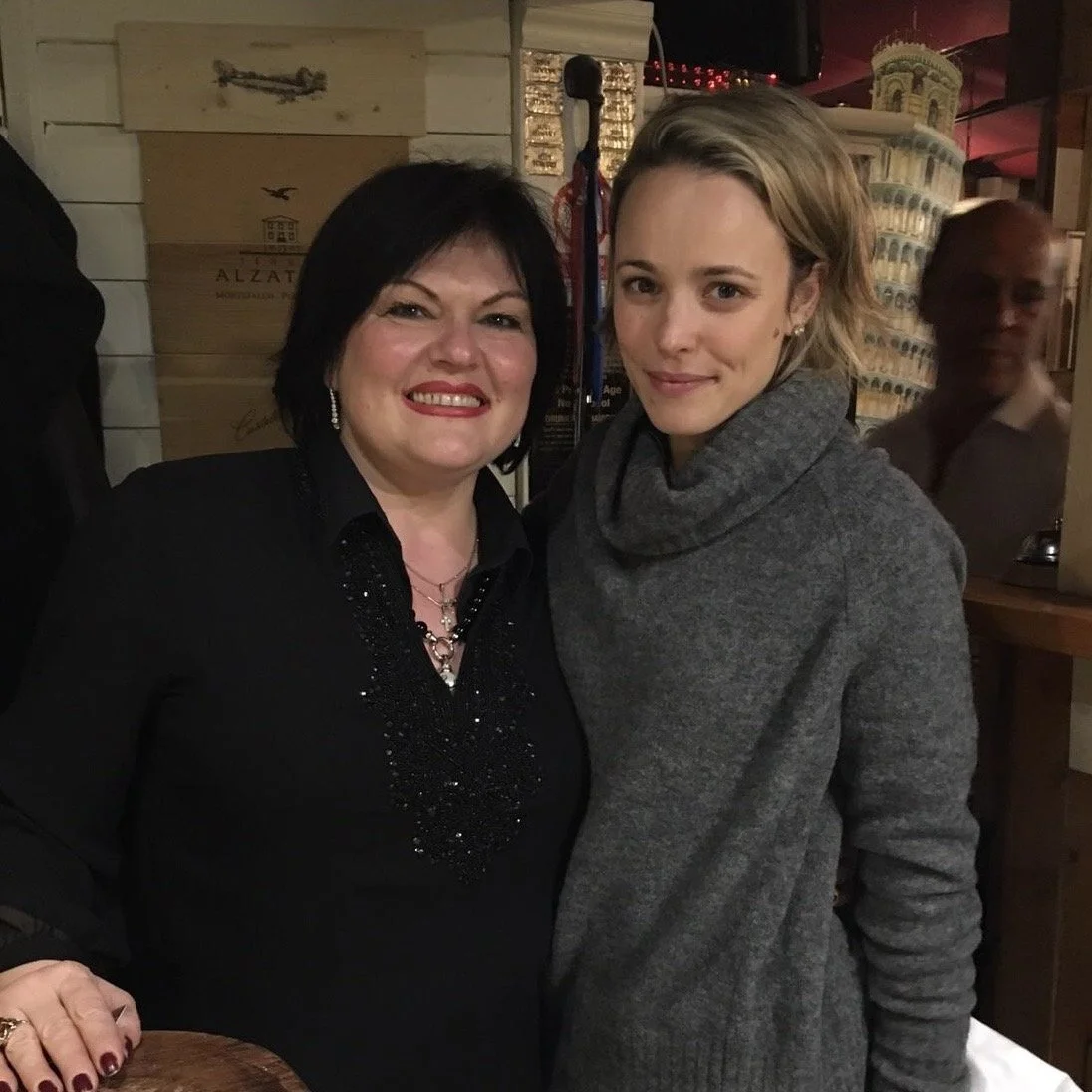 Two women smiling and standing close together in a restaurant or bar. One woman has short black hair, wears a black top with embellishments, and jewelry. The other woman has short blonde hair, wears a gray turtleneck sweater. Wooden crates and decora