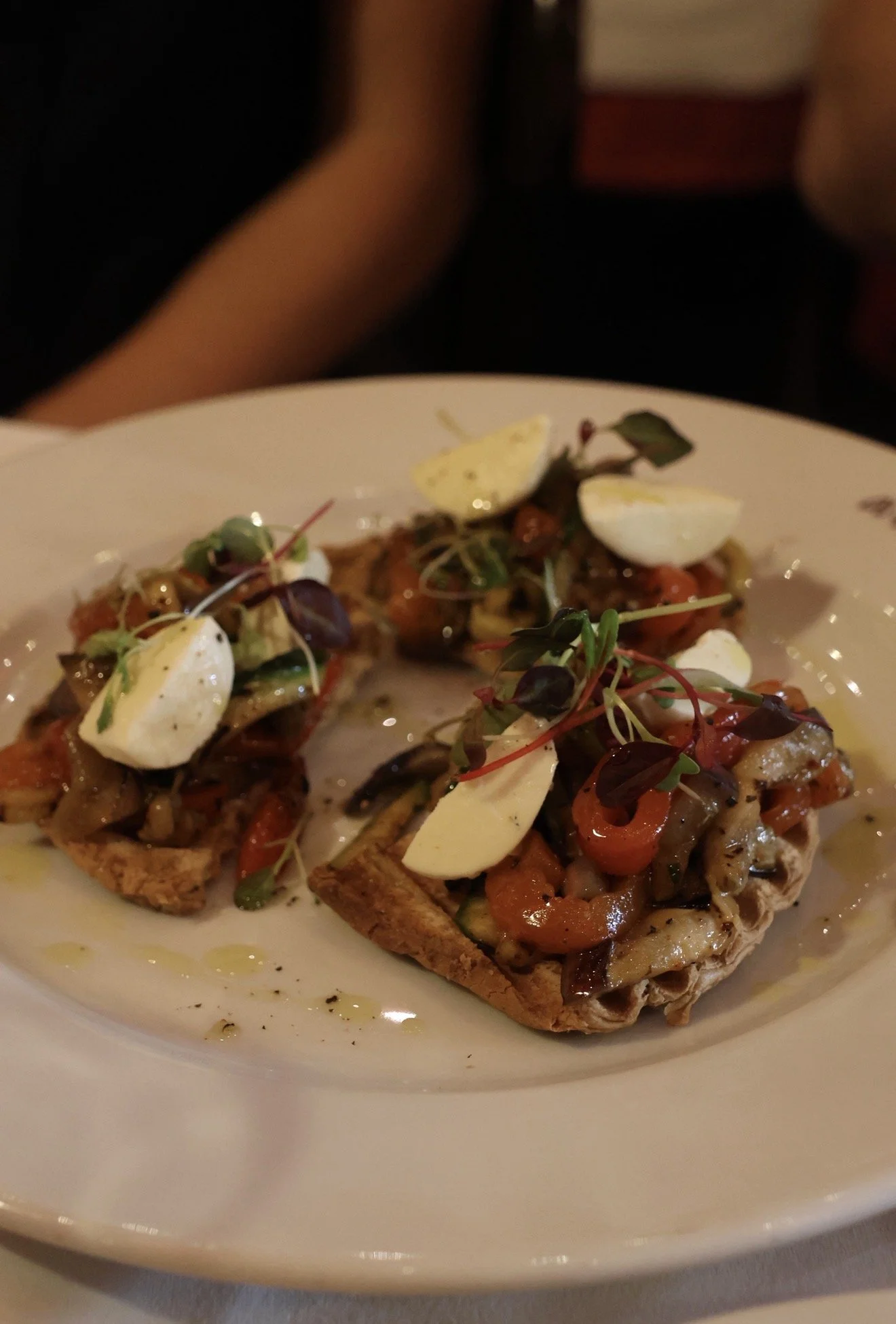 A close-up of a plate of toasted bread topped with roasted vegetables, dollops of cheese, and microgreens, drizzled with olive oil.