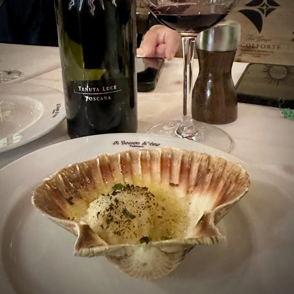 A dish served in a large shell with melted cheese and herbs, placed on a white plate at a restaurant. In the background, there is a bottle of red wine, a glass of red wine, a wooden salt shaker, and a person using a smartphone.
