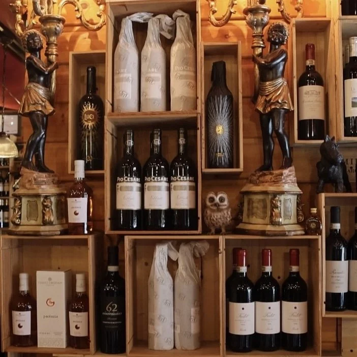 Shelf display with wine bottles, decorative owl figure, wooden sculptures of children, and wood-paneled background.