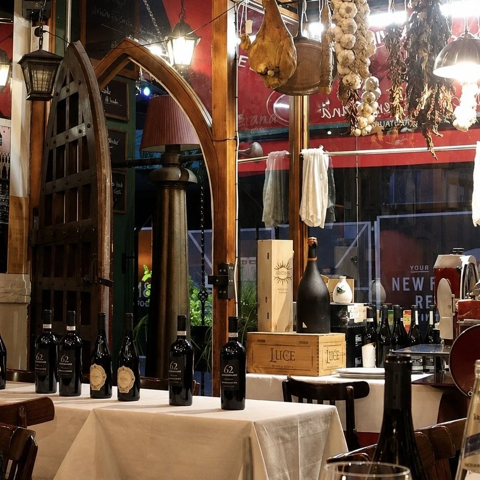Interior of a cozy restaurant or wine bar with a long tied table, bottles of wine, garlic hanging from the ceiling, and decorative items like a lamp, wooden crates, and dried herbs.