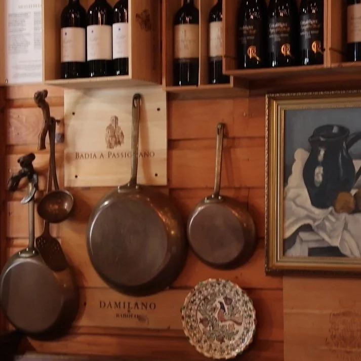 Wall decorated with hanging cast iron pans, a framed painting of a pitcher and a dish, bottles of wine, a wooden box with a logo, and a decorative plate.