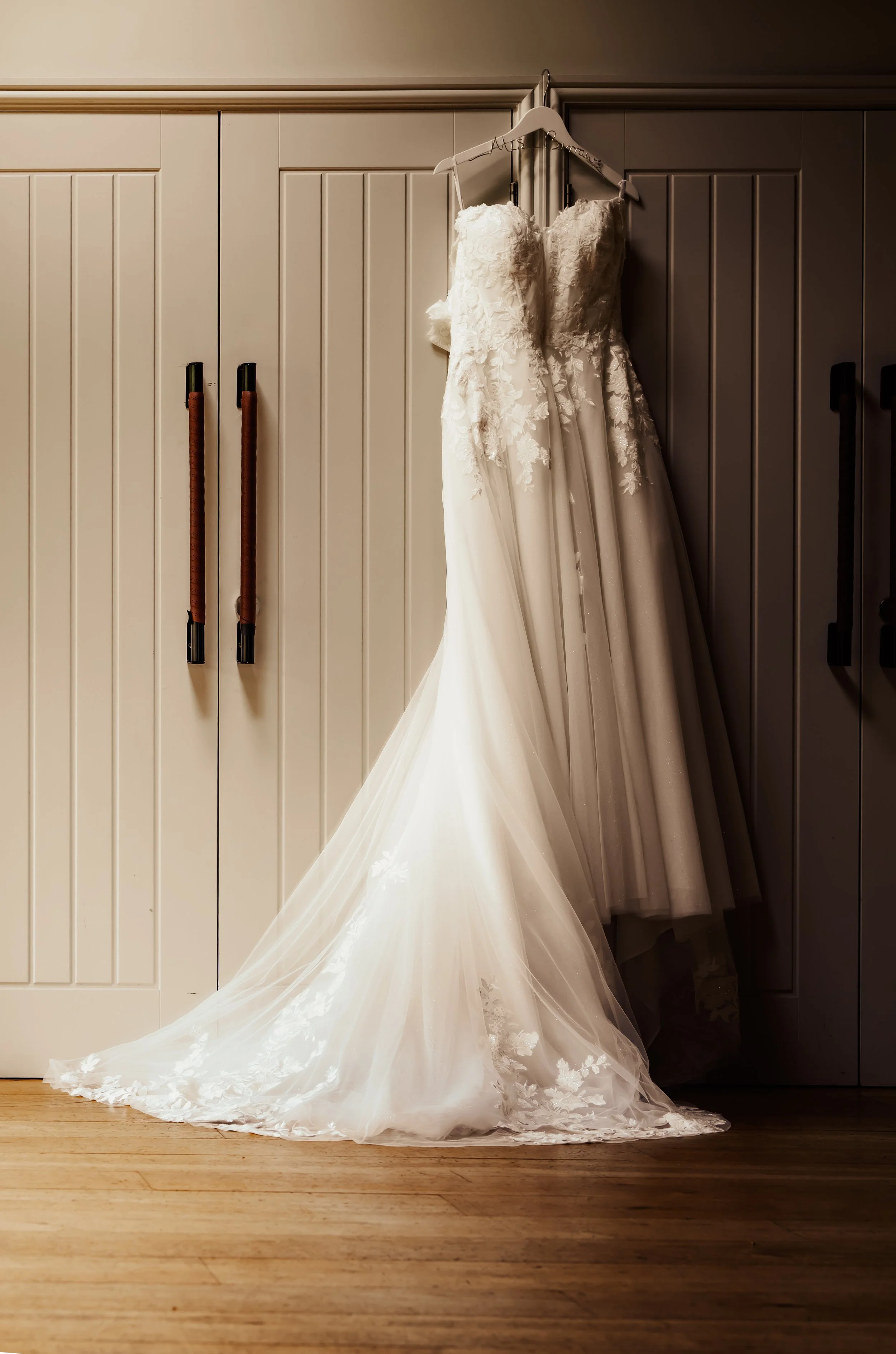 Wedding dress hanging on a white hanger on a closet door, with a long train spreading onto the wooden floor.