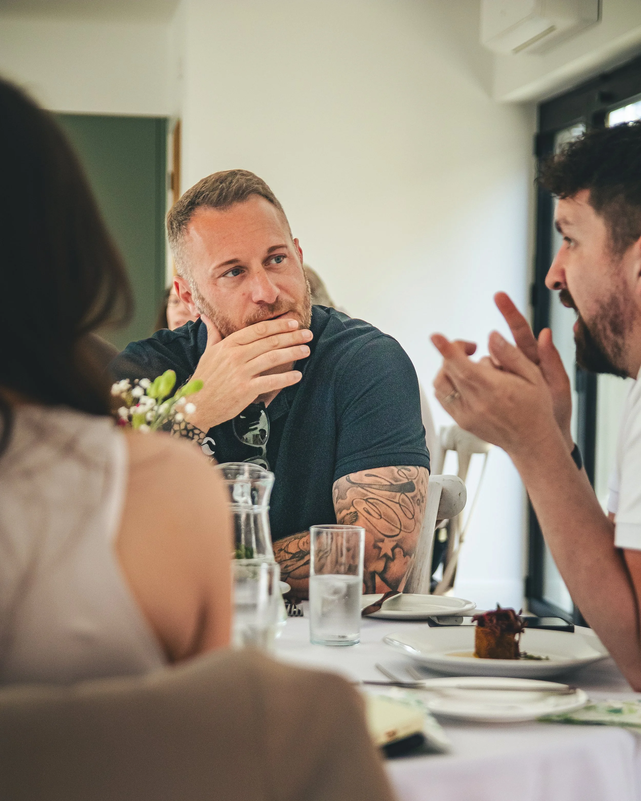 Two men are engaged in a conversation at a table during a meal, with a cake and drinks on the table, inside a bright room with white walls and large windows.