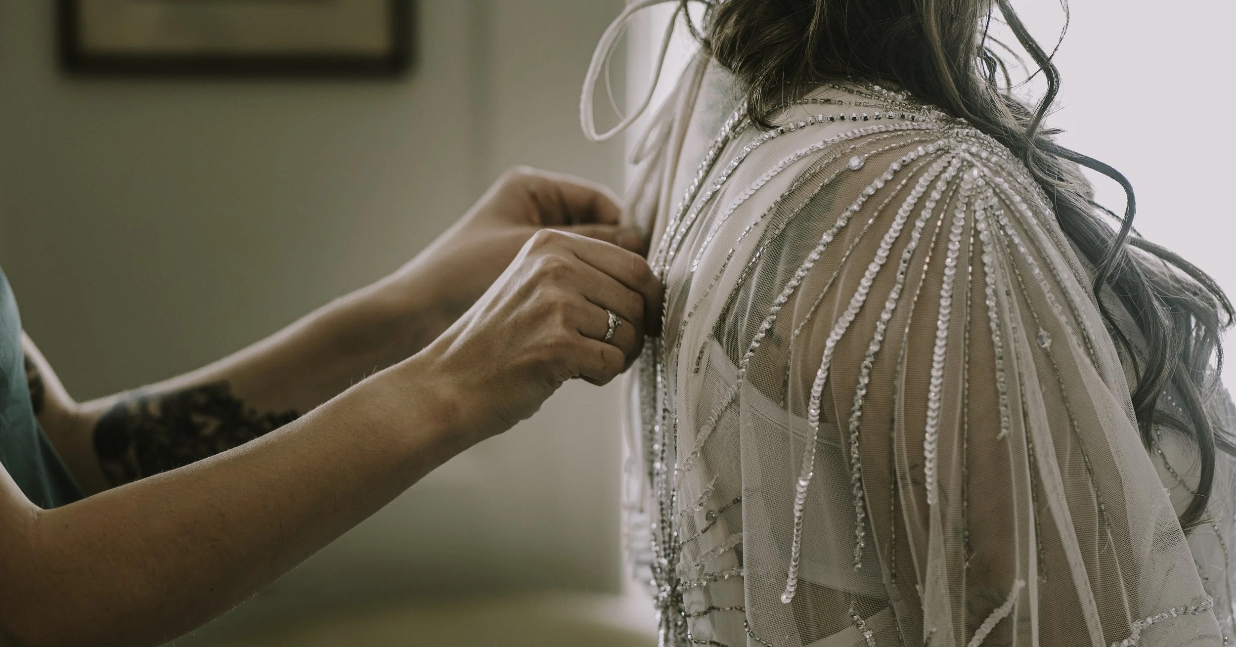 A woman is helping a bride with her wedding dress, adjusting the shoulder area decorated with shiny beads and sequins.