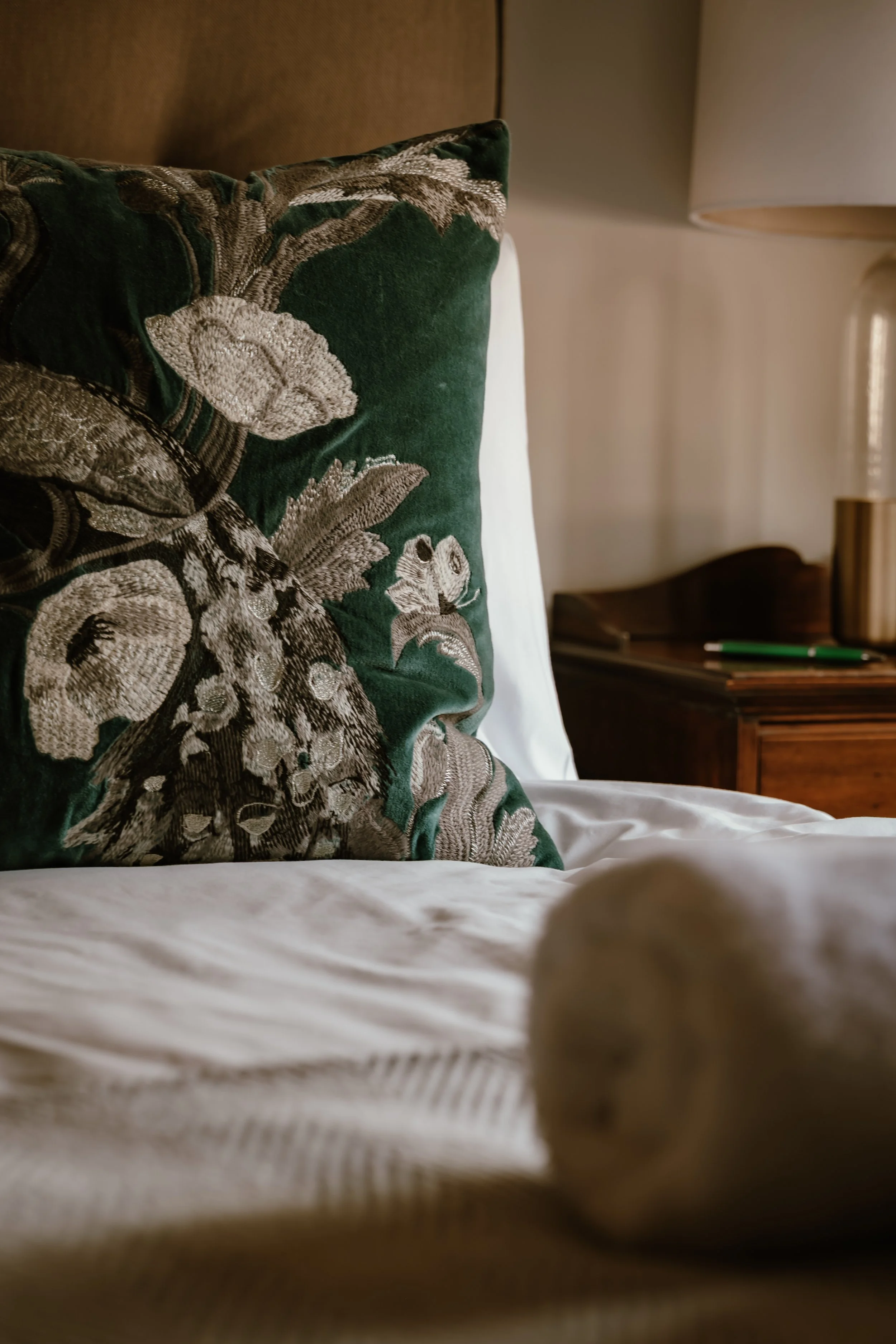 Close-up photo of a decorative pillow with a floral pattern on a bed, with a nightstand and lamp visible in the background.