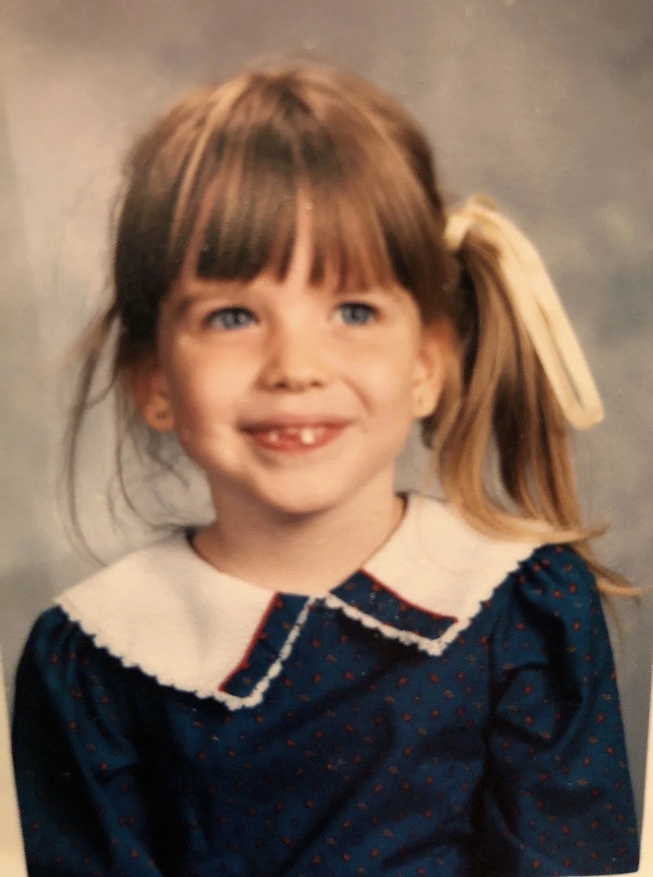 A young girl with light brown hair styled in a ponytail with a large white ribbon, smiling and showing her teeth with a gap in her front, wearing a navy blue dress with red polka dots and a white Peter Pan collar with red trim.