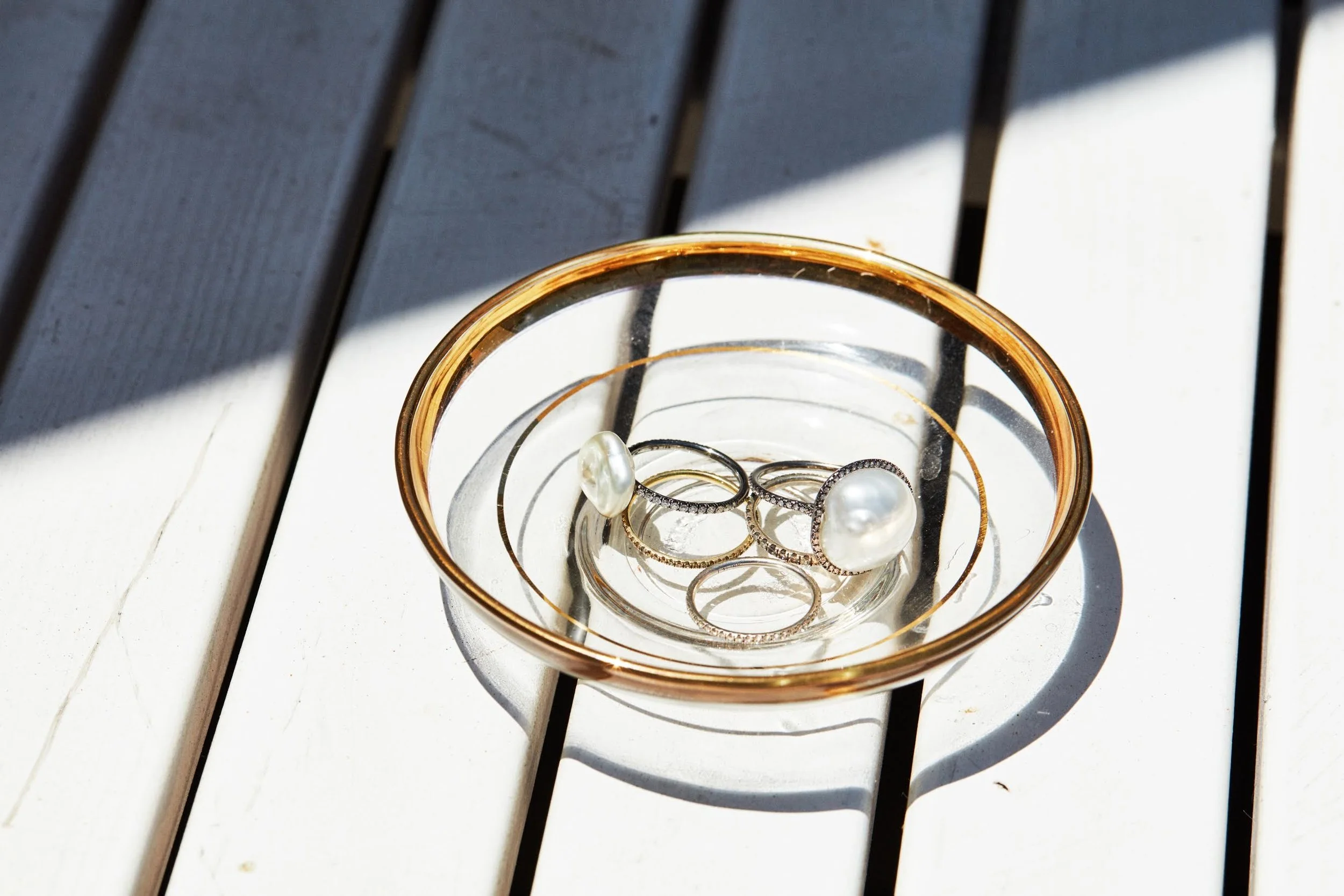 A round glass container with gold trim inside a clear glass bowl, holding pearl earrings and rings, on a white wooden surface with shadows.