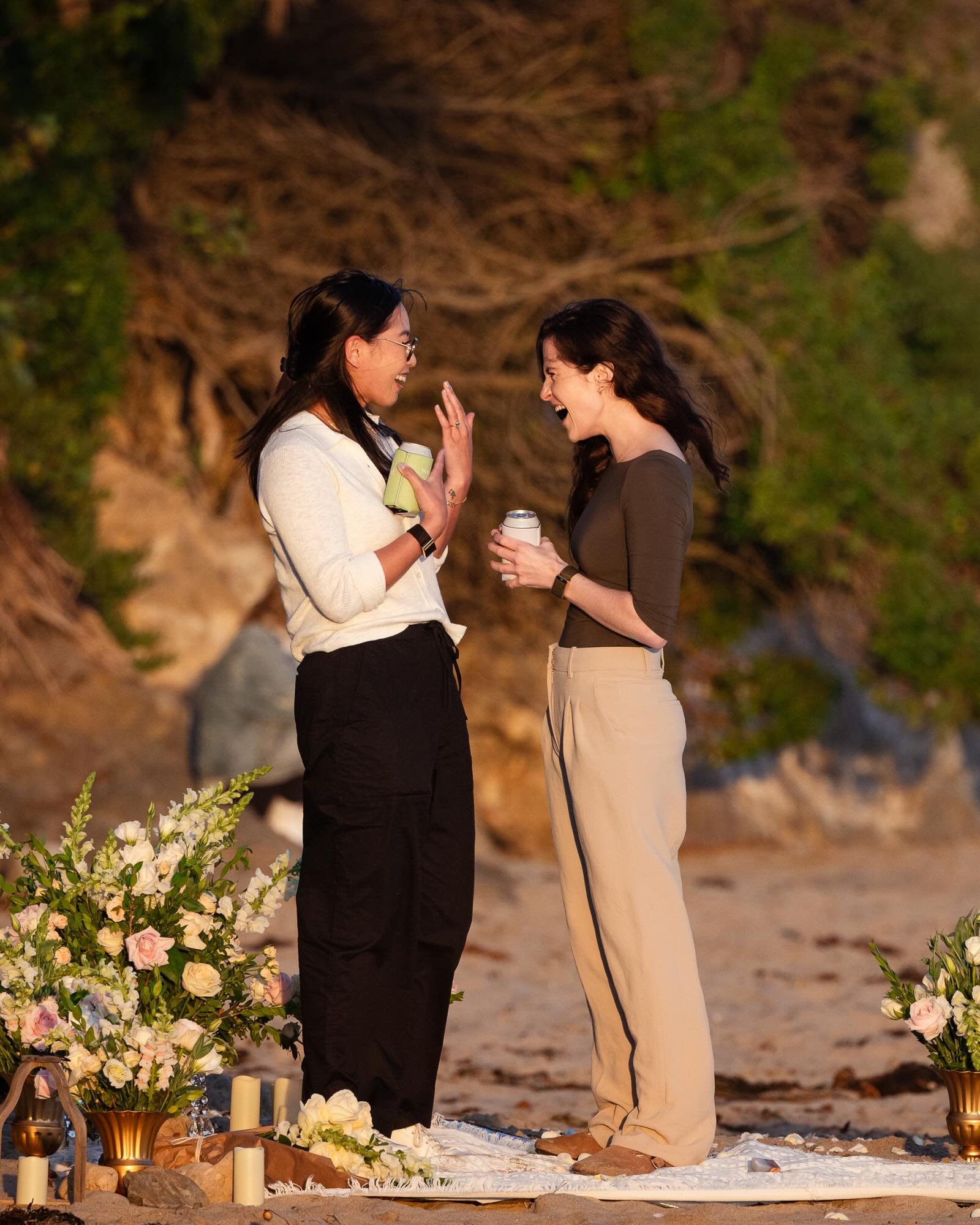 Two women standing on a beach, facing each other, smiling, and laughing during a daytime outdoor celebration or ceremony. They each hold a drink, and flower arrangements are arranged on the ground nearby.