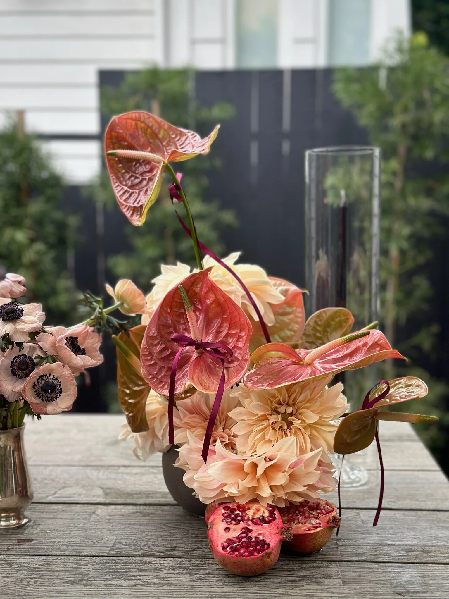 A floral arrangement with peach, pink, and red flowers and an anthurium with pinkish-red leaves and a yellow spadix, complemented by pomegranate halves in front of it, placed on a wooden table with a blurred outdoor background.