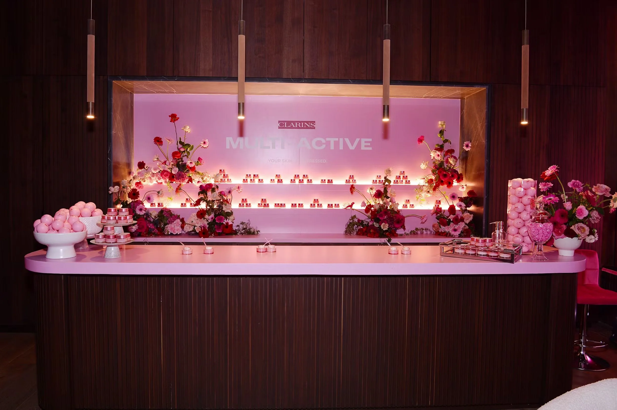 Display table with pink and white skincare products and flowers in a boutique setting, with a pink sign in the background reading "Clarins Multi-Active".