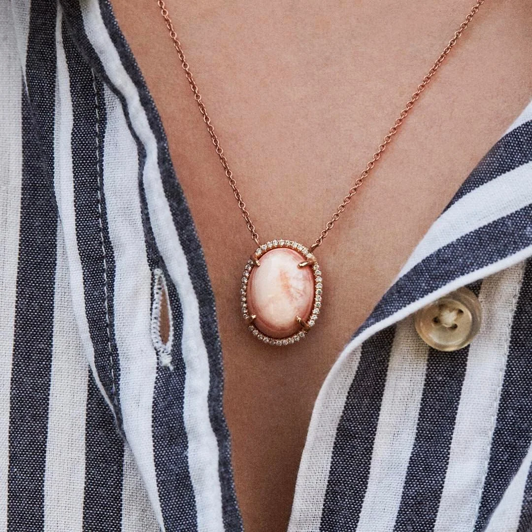 A necklace with an oval peach-colored gemstone surrounded by small diamonds, worn by a person dressed in a black and white striped shirt.