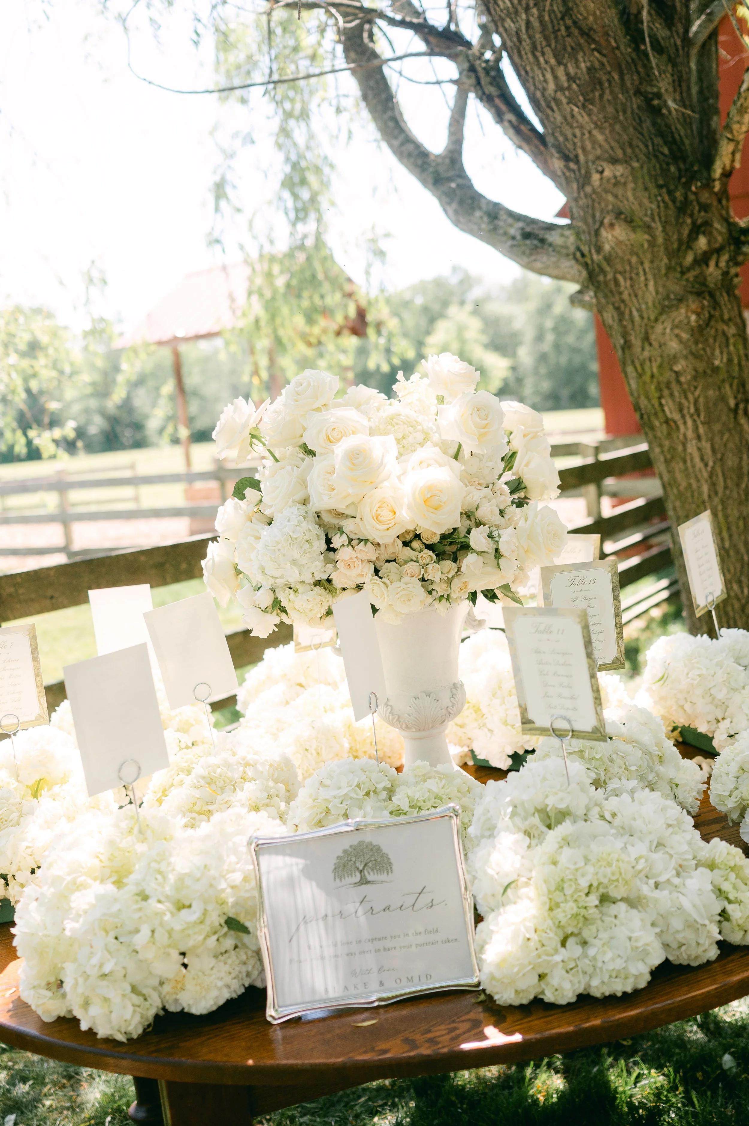 A bouquet of white roses and hydrangeas arranged in a white urn on a wooden table, surrounded by small white floral arrangements and display cards, outdoors with trees and a wooden fence in the background.