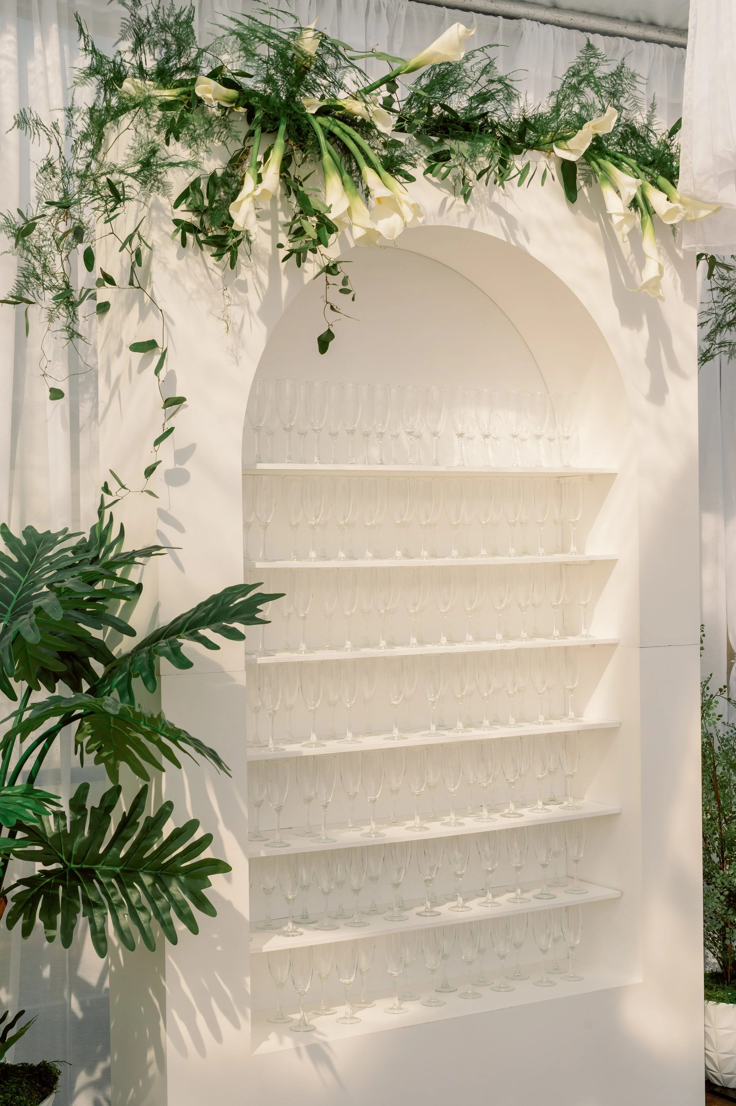White shelving unit with multiple levels holding evenly spaced empty wine glasses, decorated with white flowers and greenery on top, and surrounded by various green plants.