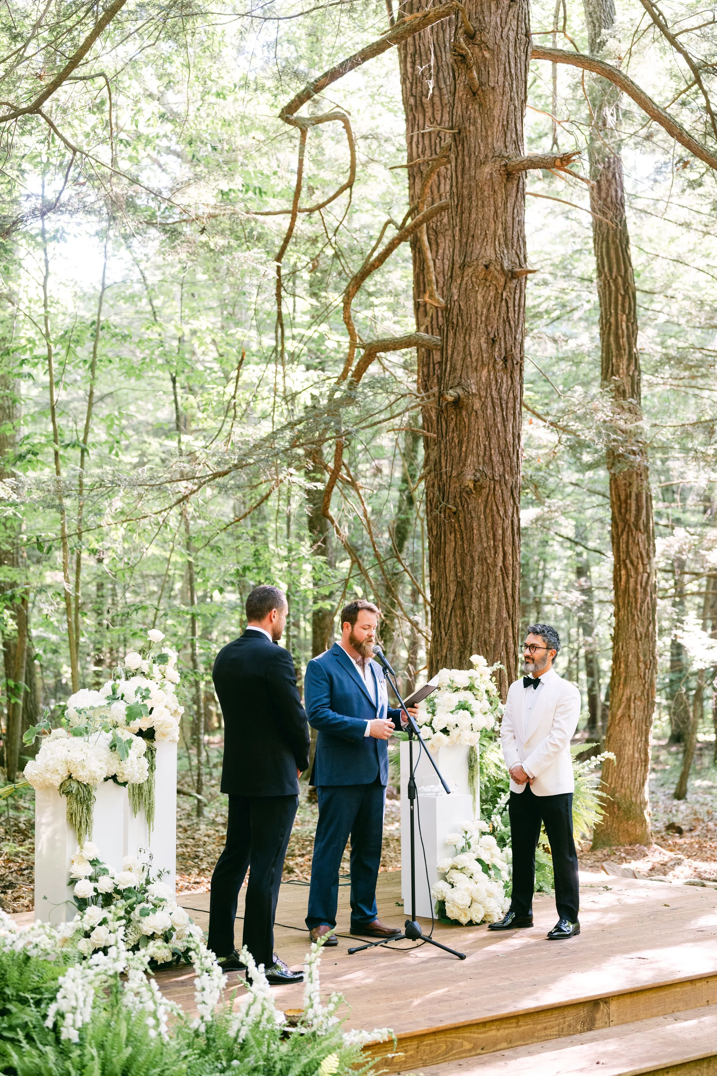 A wedding ceremony taking place outdoors in a forest with tall trees and natural sunlight. Four men are participating, with one man reading from a book at a microphone, and the others listening attentively. The scene is decorated with white flowers and greenery on white pedestals surrounding the ceremony area.