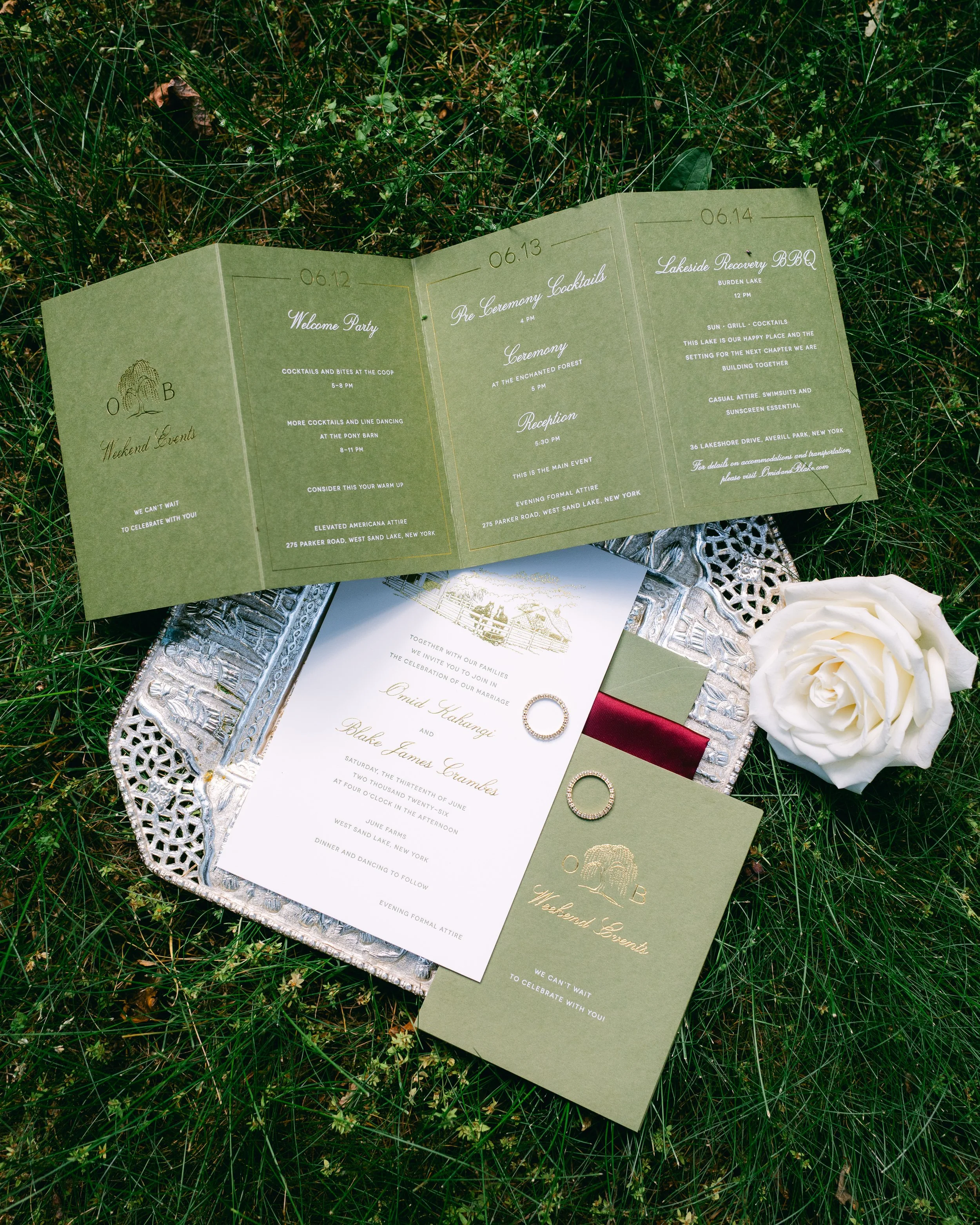 Wedding invitation suite on a grassy surface with white roses, silver tray, and rings.