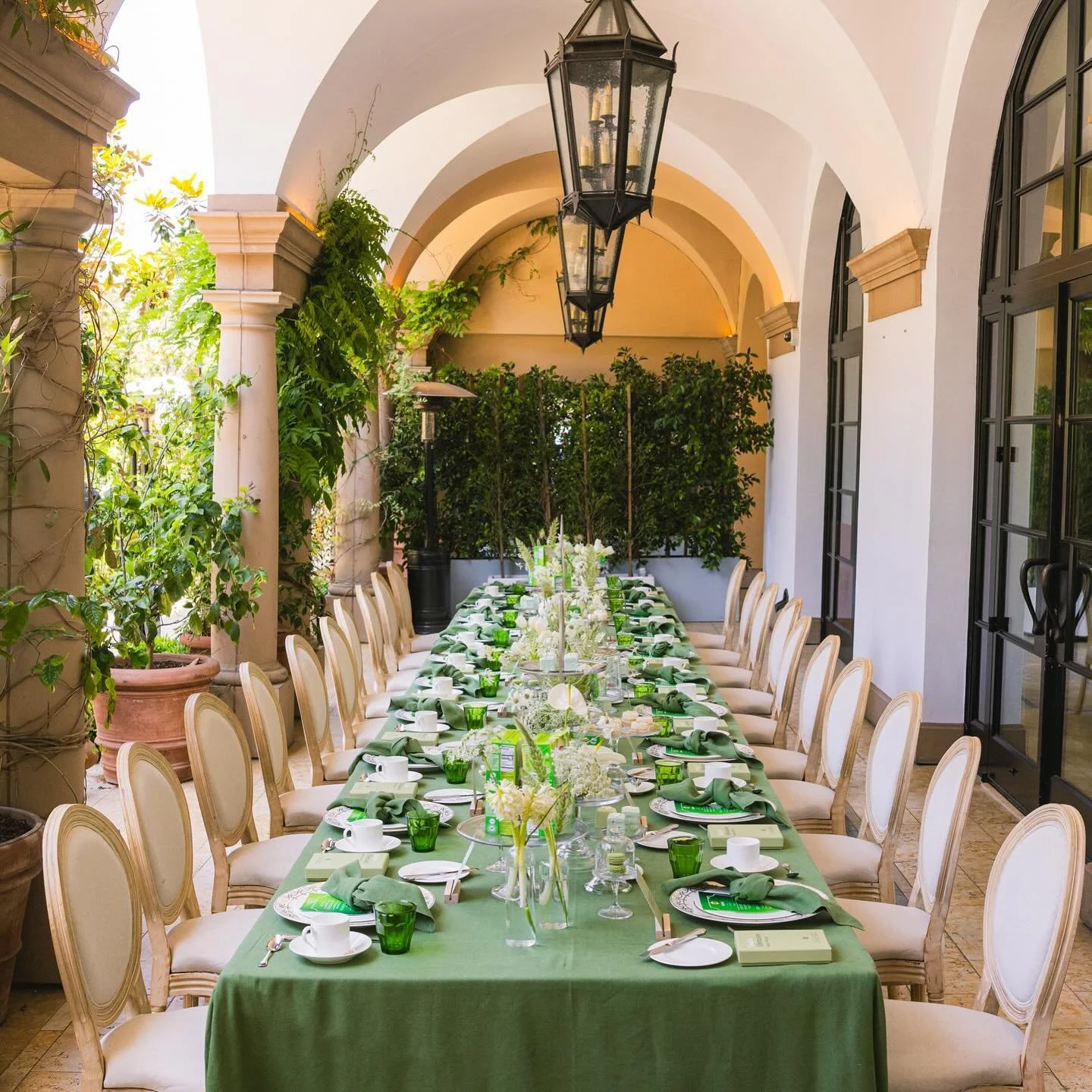 Long dining table set for a meal in an outdoor covered patio with white chairs, green tablecloth, and floral centerpieces, surrounded by plants and large windows.