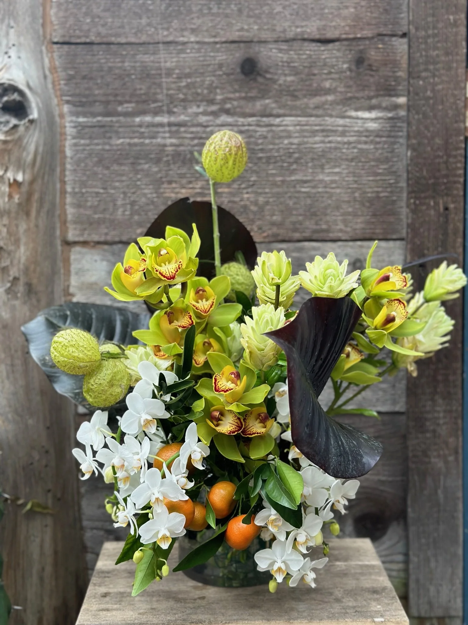 A floral arrangement with yellow orchids, white orchids, orange fruits, and dark purple leaves against a wooden background.