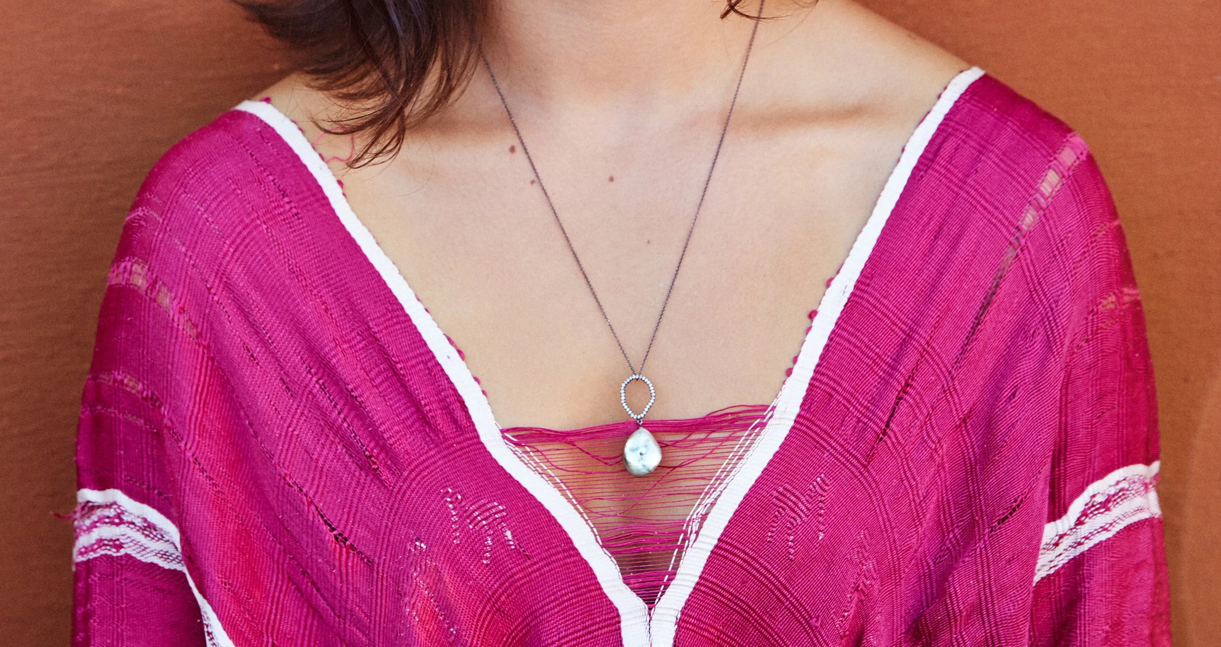 Close-up of a person's upper torso wearing a pink, semi-transparent, knitted top with white trim and a layered silver necklace with a teardrop-shaped pearl pendant.
