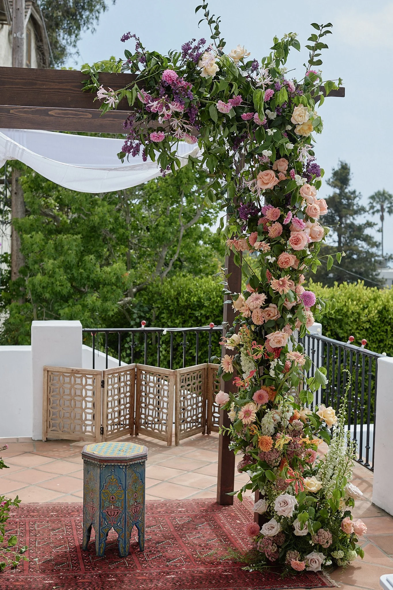 A decorated outdoor area with a flower garland and a small, ornate stool on a rug, featuring a wooden screen, a terracotta tiled floor, and trees in the background.