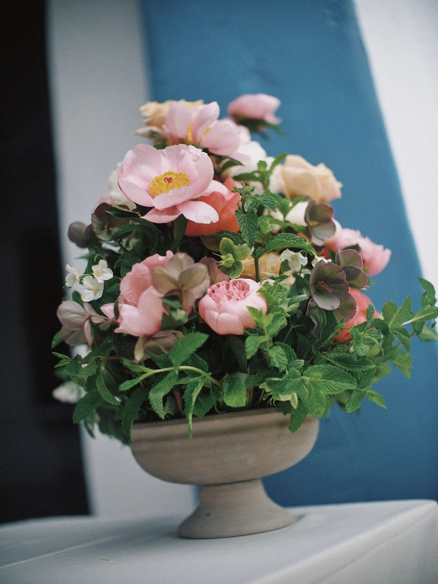 A beige decorative vase filled with pink, white, and green artificial flowers and green foliage, placed on a white surface against a blue background.