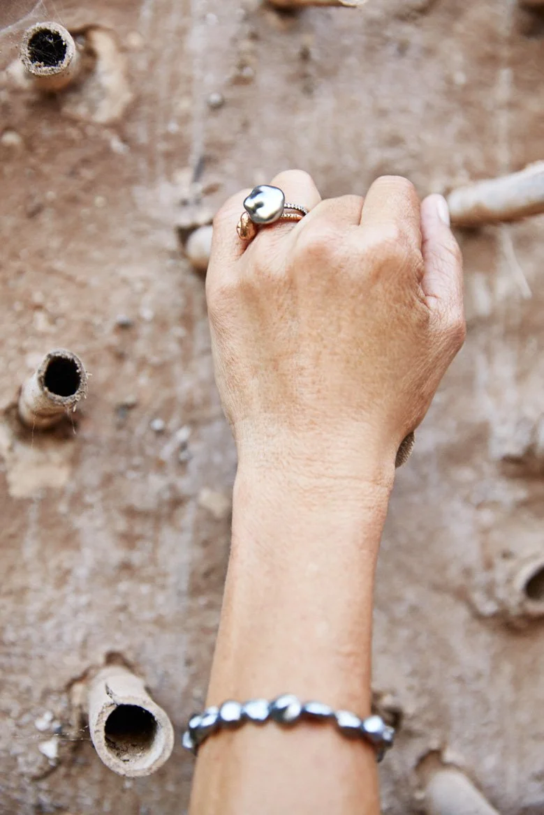 A person's hand with a pearl ring and a pearl bracelet, reaching up toward a textured dirt wall with holes.