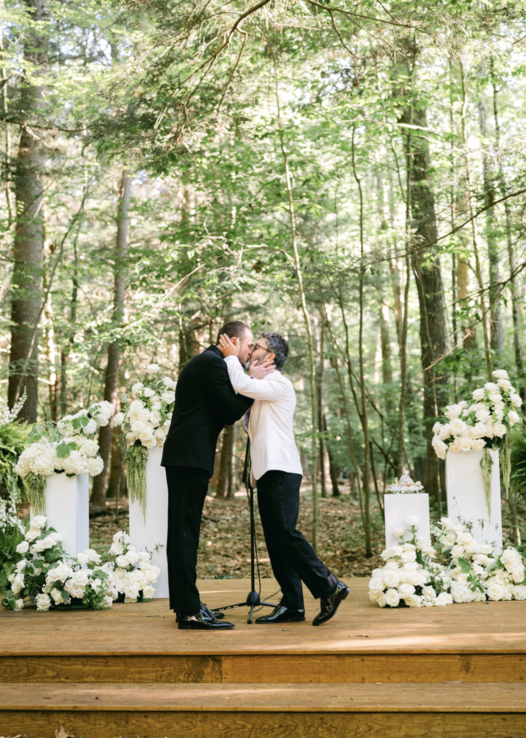 Two men in formal attire sharing a kiss at an outdoor wedding ceremony on a wooden platform, surrounded by white floral arrangements and greenery.