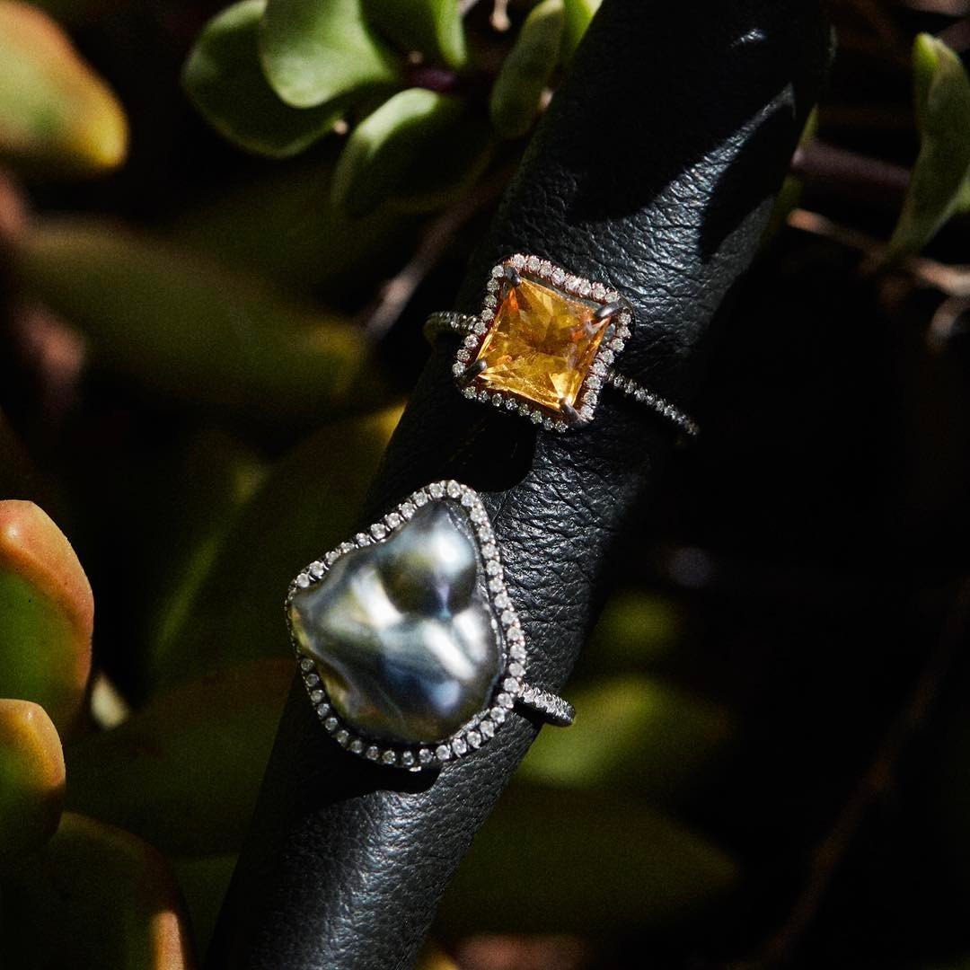 Close-up of two rings on a black leather surface surrounded by green leaves. One ring has a large, square yellow gemstone surrounded by small diamonds. The other ring features a baroque pearl with an irregular shape, also bordered by small diamonds.