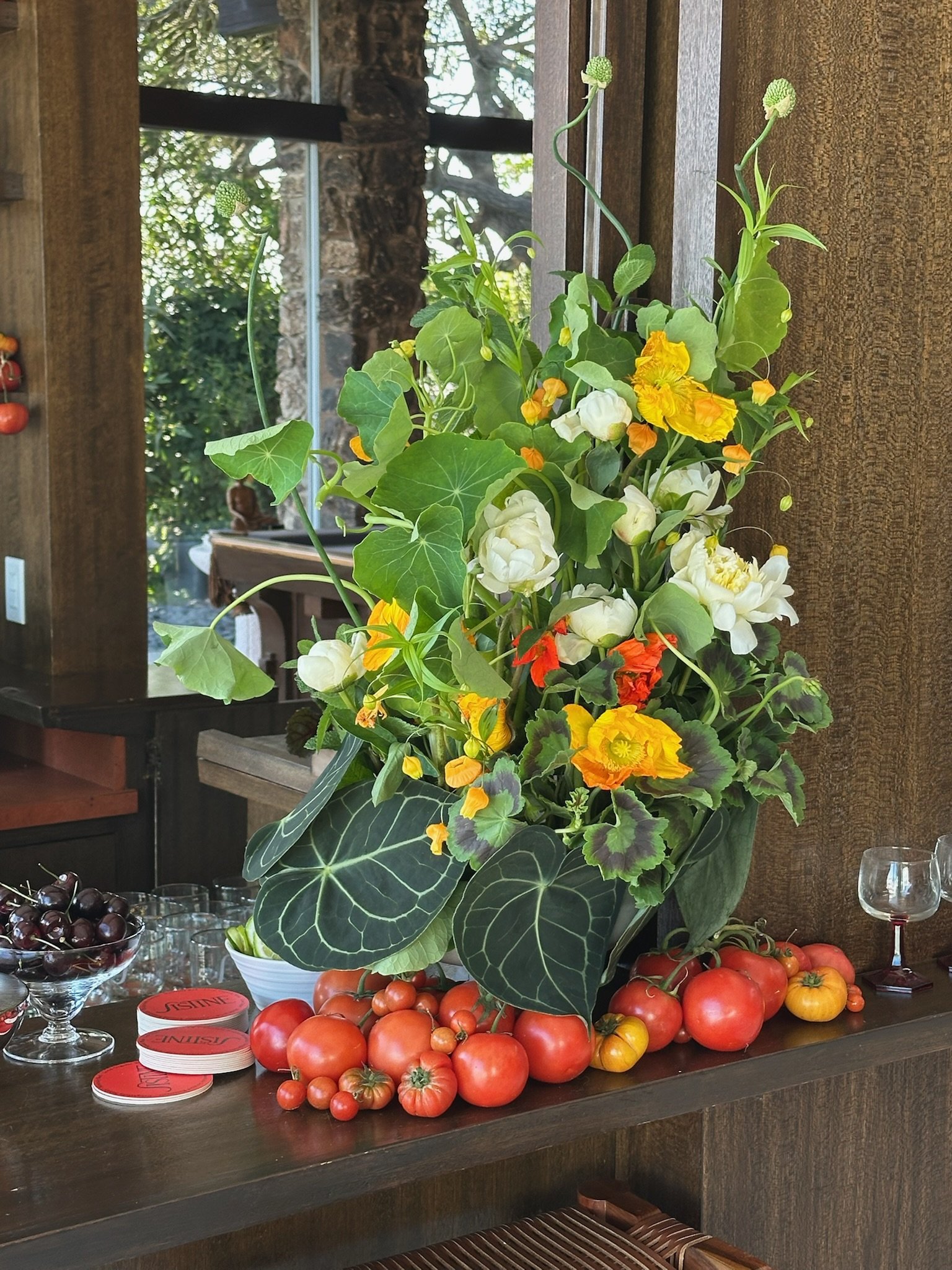 A large floral arrangement with green leaves and orange, yellow, and white flowers is placed on a wooden table. Surrounding the arrangement are tomatoes, grapes, and small containers, with a rustic wooden wall and window in the background.