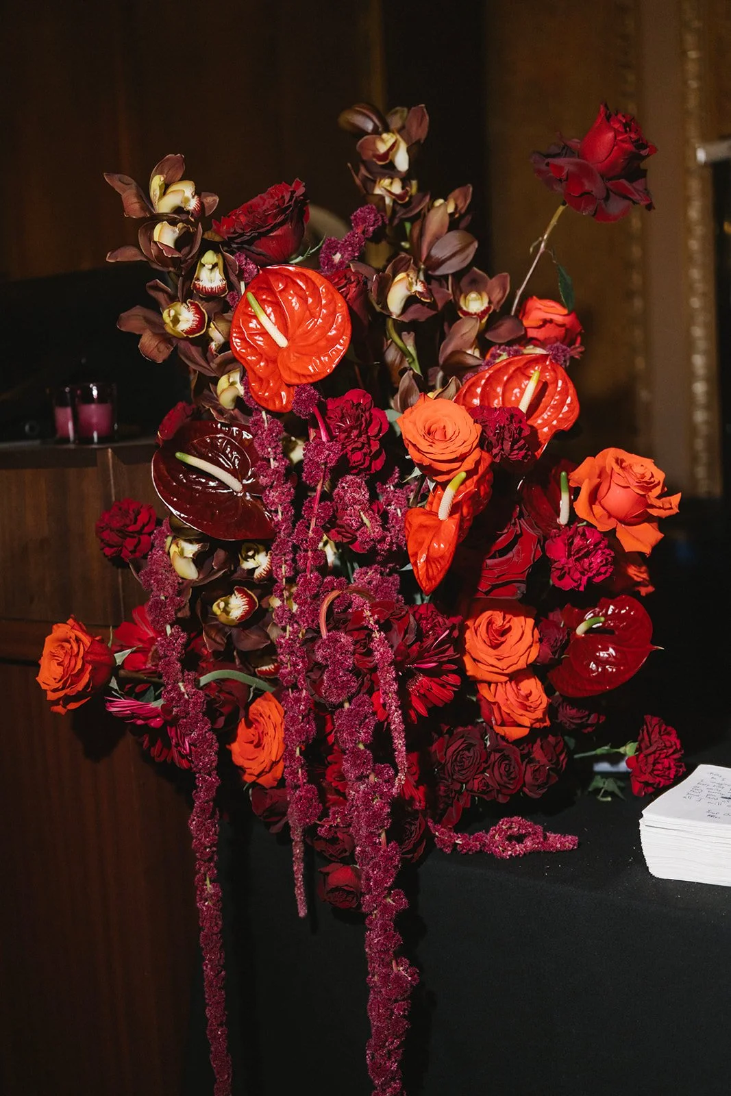 A floral arrangement with red and orange roses, dark purple orchids, red anthuriums, and pink hanging amaranthus on a black surface, with a dark background.