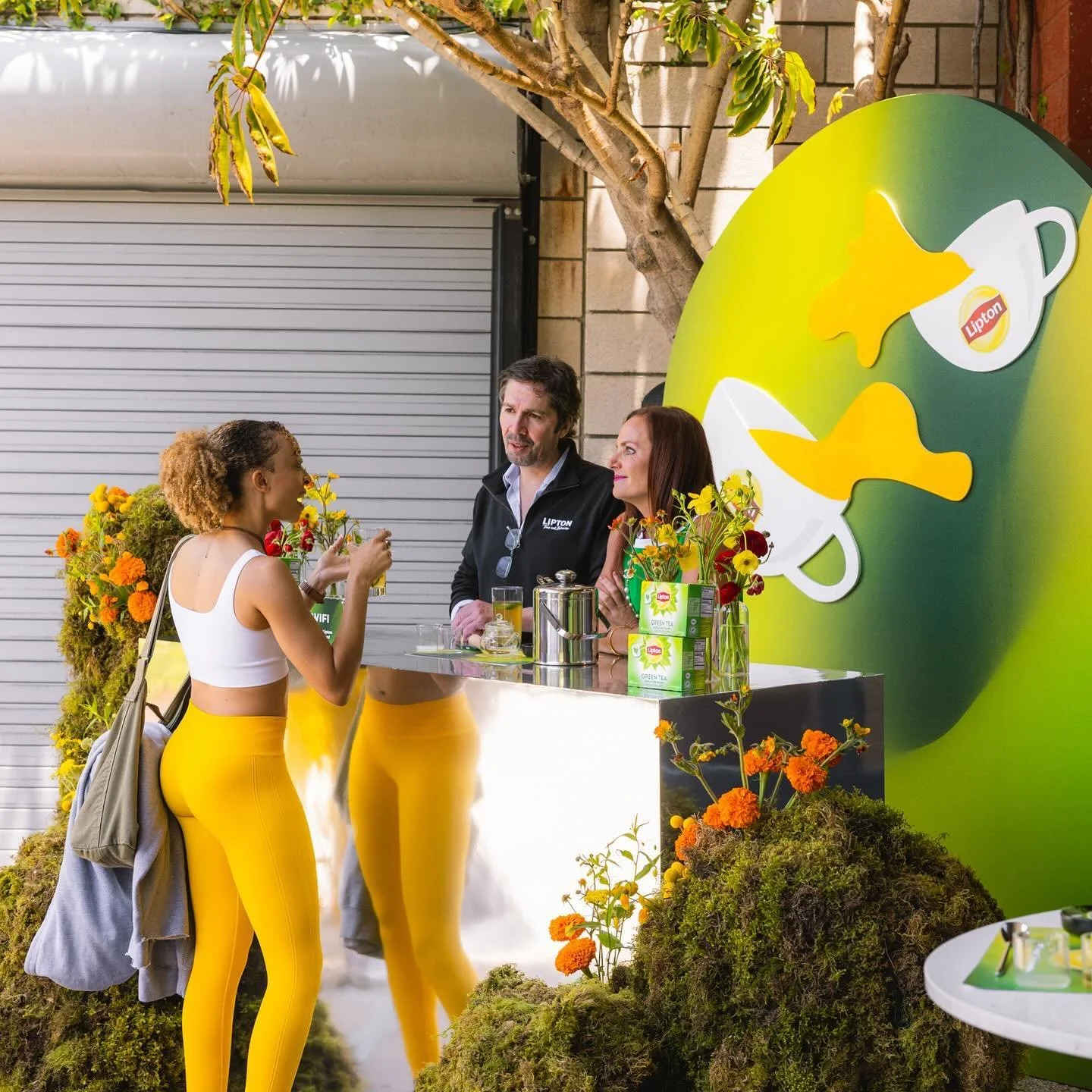 A woman in a white crop top and yellow leggings talking to two people behind a booth decorated with flowers and a green Lipton tea sign, with a large Lipton cup design in the background.