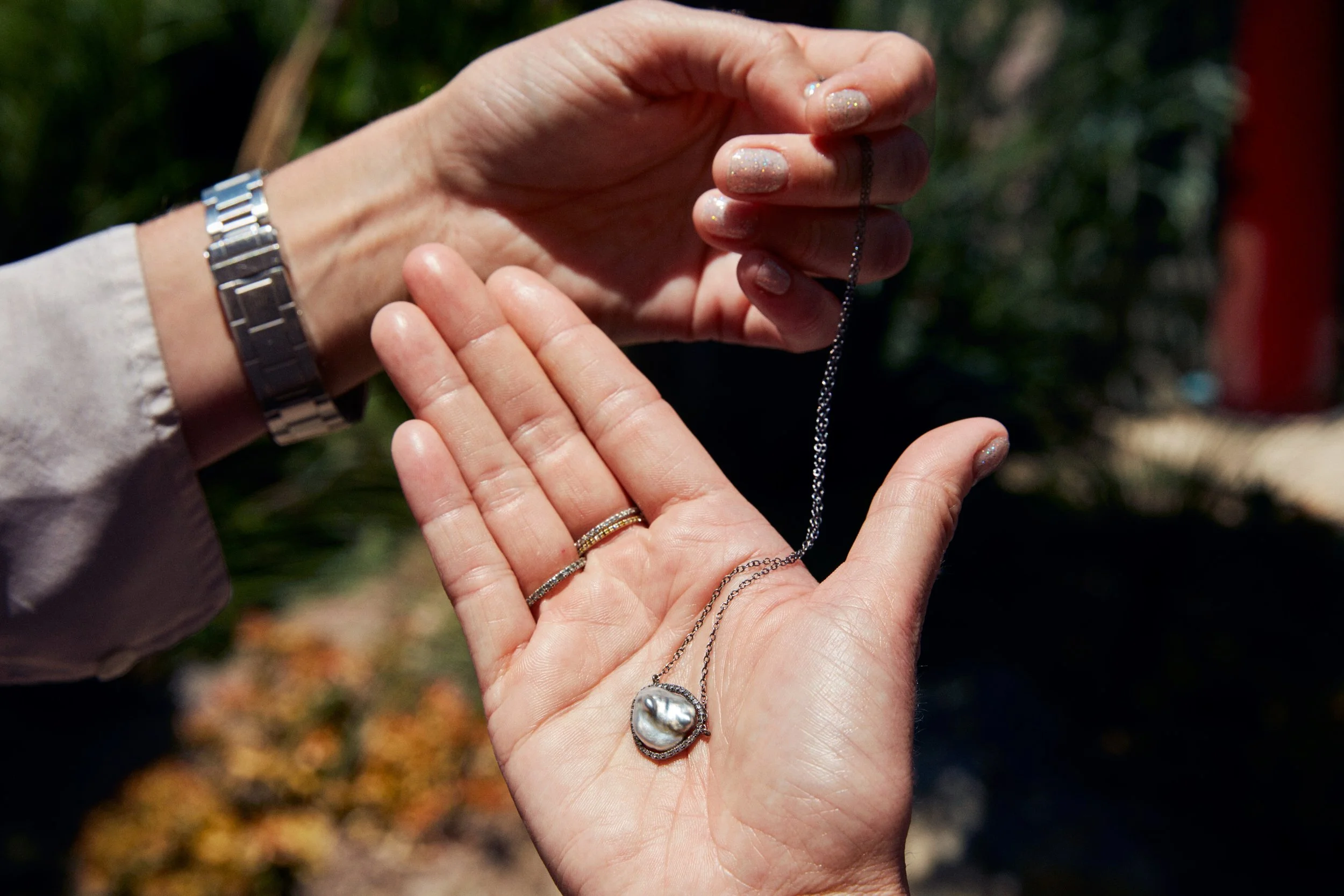 A person holding a necklace with a large pearl pendant and wearing multiple rings and a watch. The person's fingernails are decorated with sparkly nail polish. The background is blurred outdoors with greenery and autumn leaves.