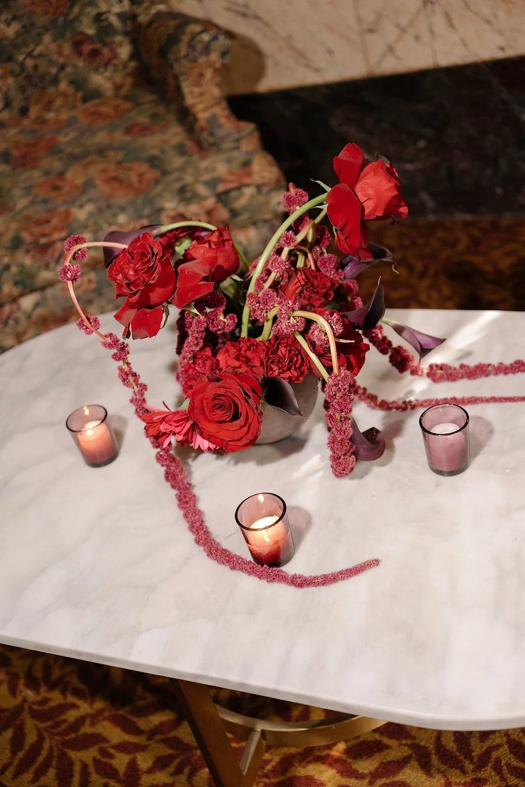A floral centerpiece with red roses, purple calla lilies, and pink accents on a white tablecloth, surrounded by four pink candle holders with lit candles, set on a round table.