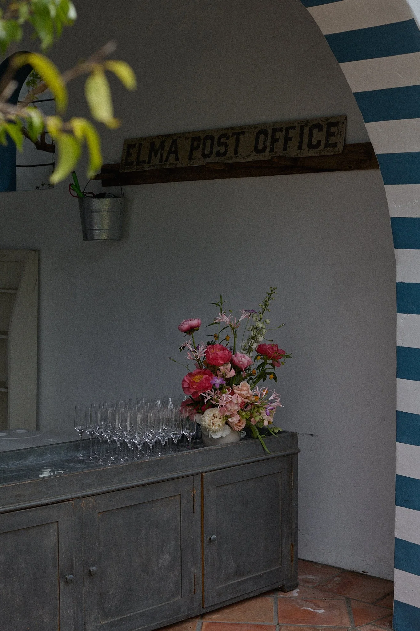 A rustic wooden sideboard with a bouquet of pink and white flowers, and empty champagne glasses arranged on it. In the background, there is a sign that reads 'Elma Post Office' and a blue and white striped wall.