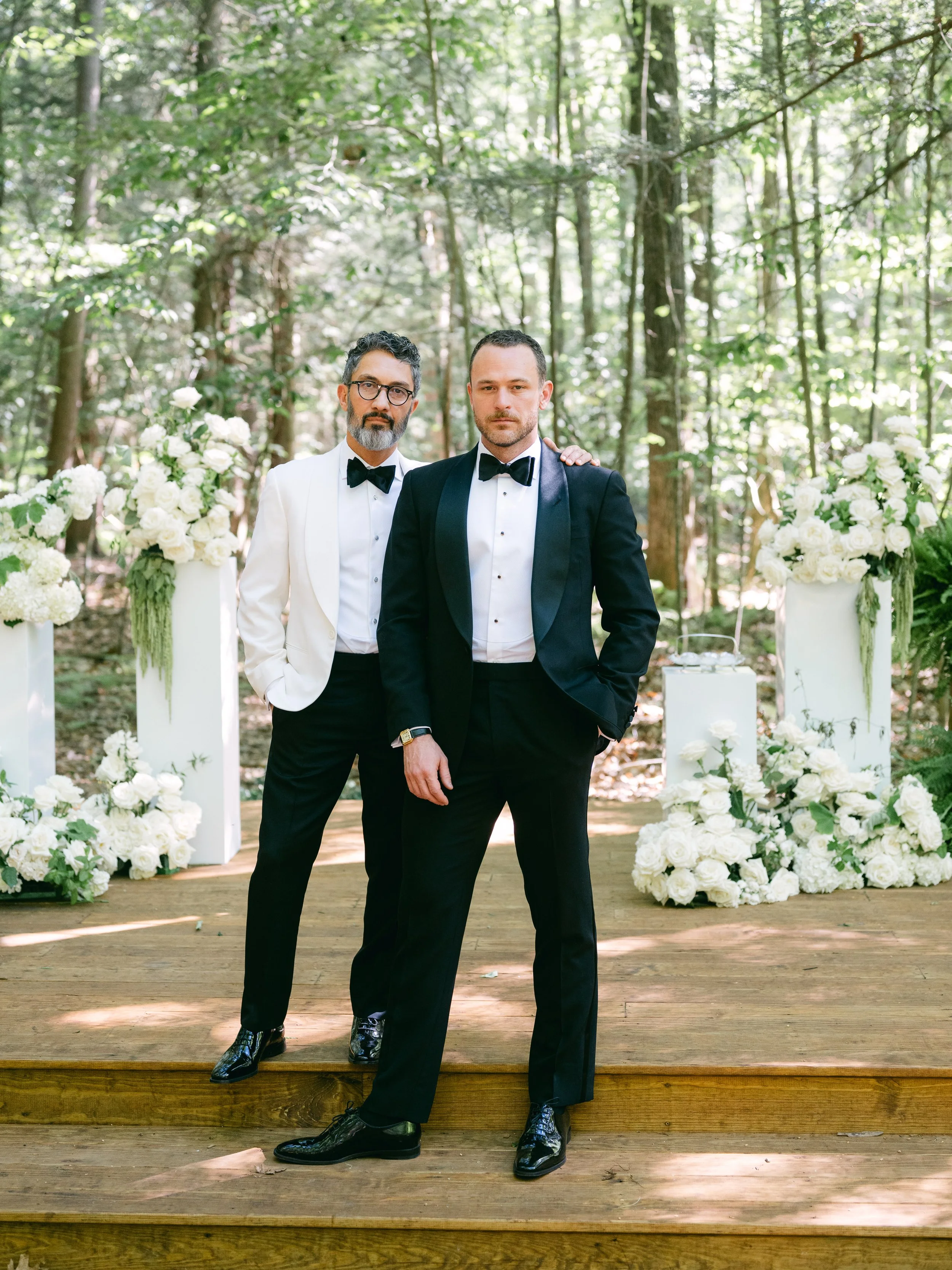 Two men in tuxedos standing next to each other on a wooden platform in a forest setting, with floral arrangements behind them.