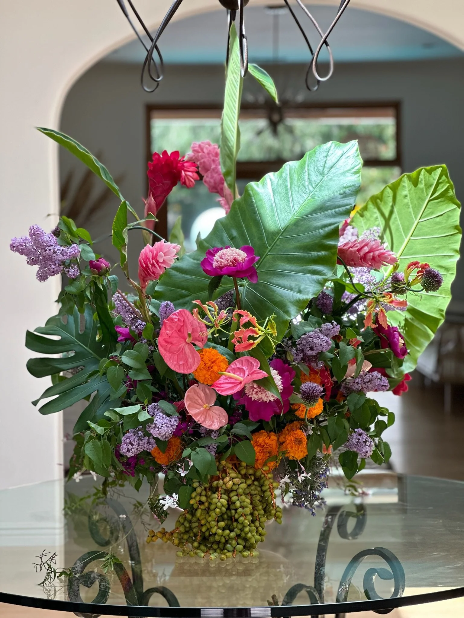 A colorful arrangement of flowers including pink, purple, and orange blossoms with large green leaves on a glass table in front of a window.