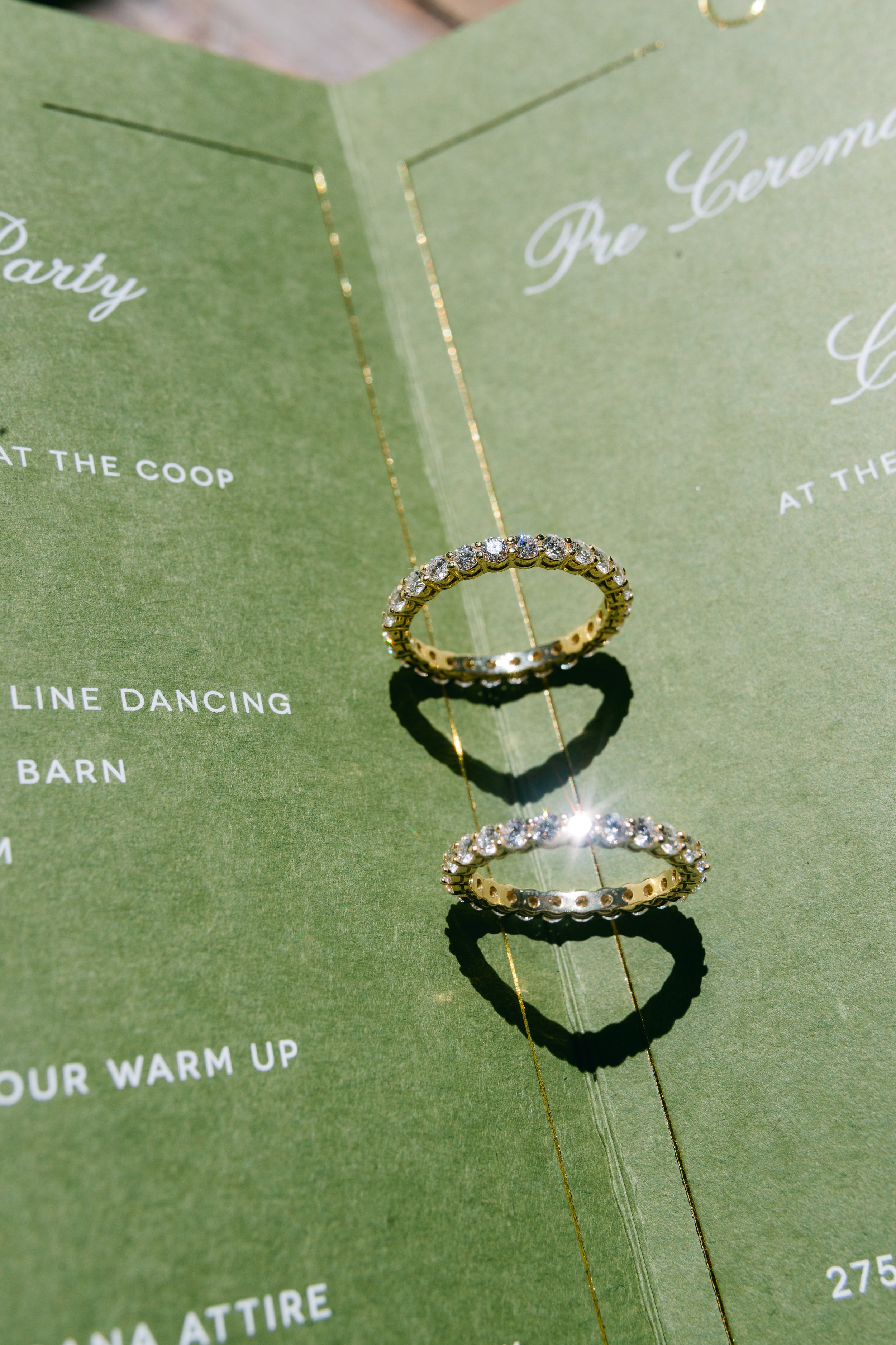 Close-up of two gold rings with diamonds placed on a green paper with white text, with one ring positioned above the other, and a gold chain necklace hanging behind them.