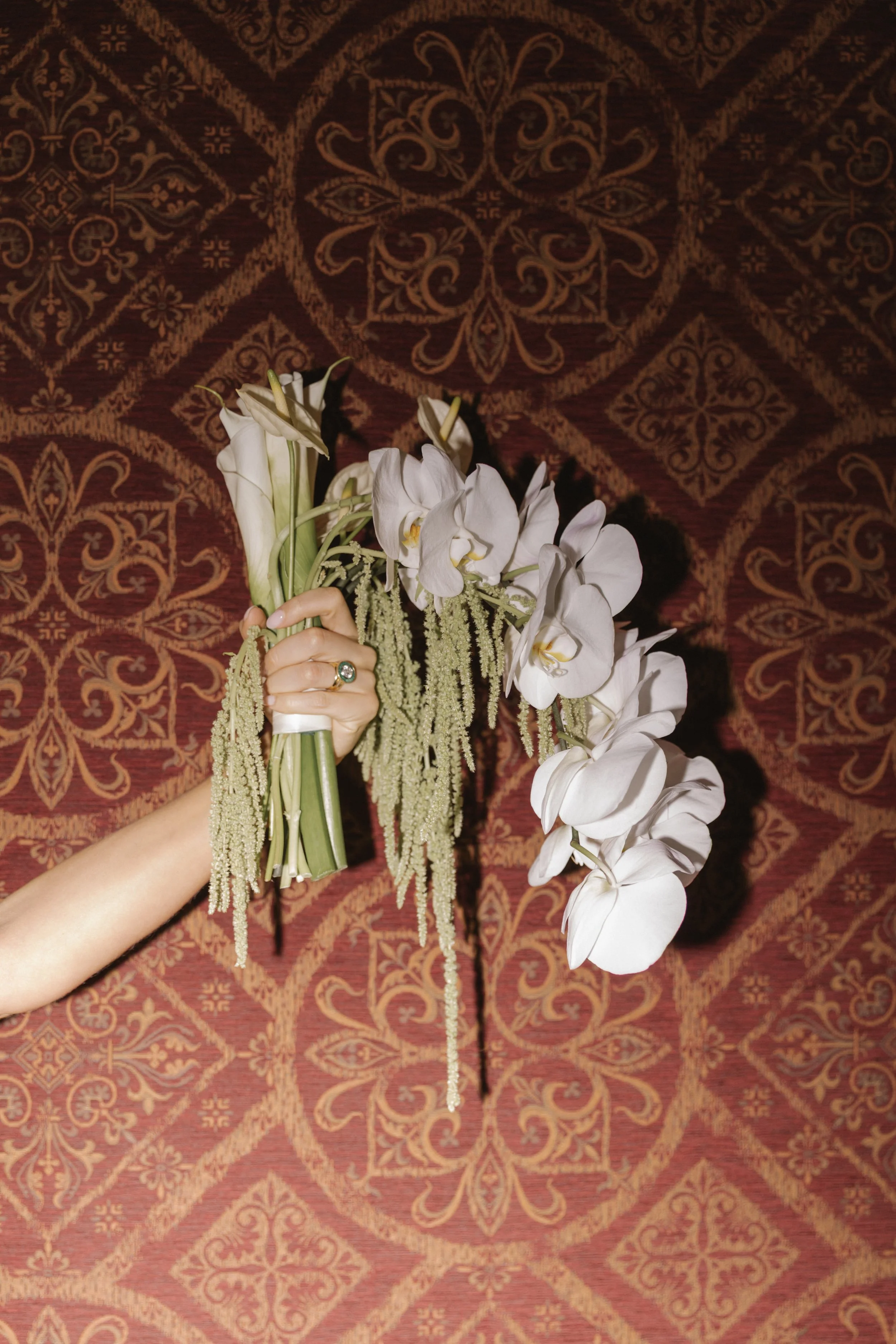 A person holding a bouquet of white flowers with greenery against a red and gold patterned carpet.